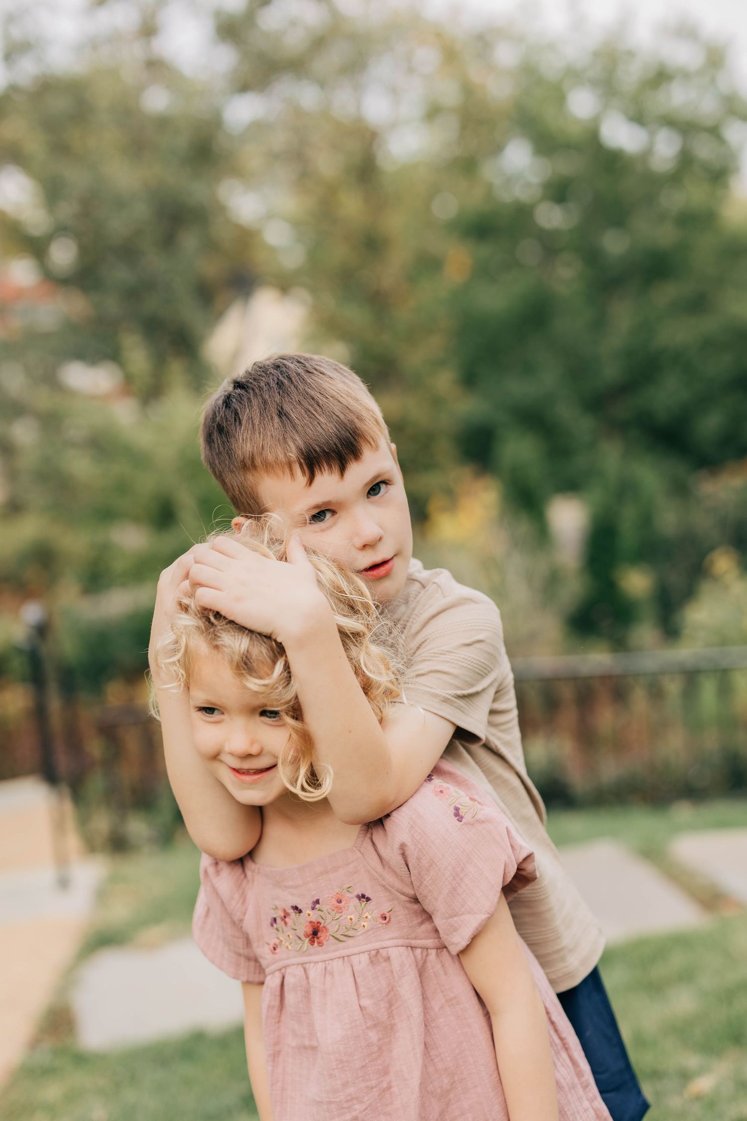 Young boy in a tan shirt hugging a smiling girl in a pink floral dress from behind on a sunny garden path.