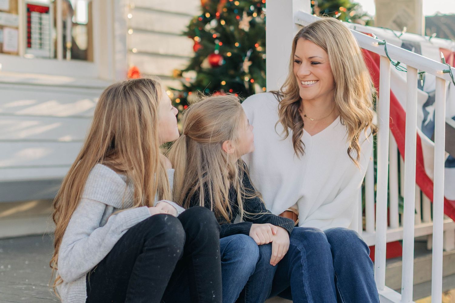 A mother and two daughters share a joyful moment on porch steps with a Christmas tree in the background.