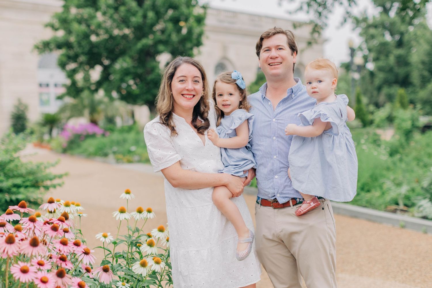 Happy family of four posing outdoors among colorful flowers, parents holding two young daughters in a garden setting.