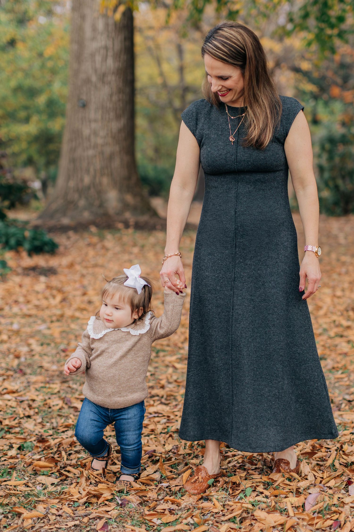 Mother holds toddler daughter's hand while walking through fallen autumn leaves in a park.