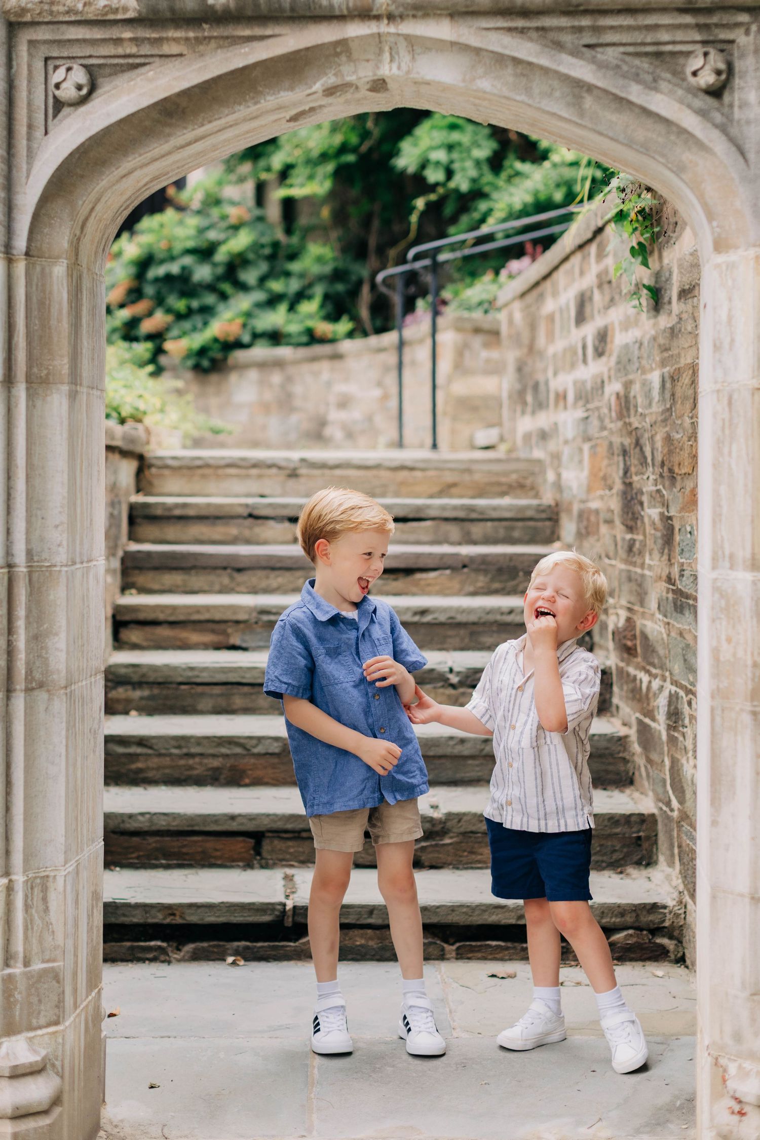 Two young blond boys laughing and playing under a stone archway with steps in the background.