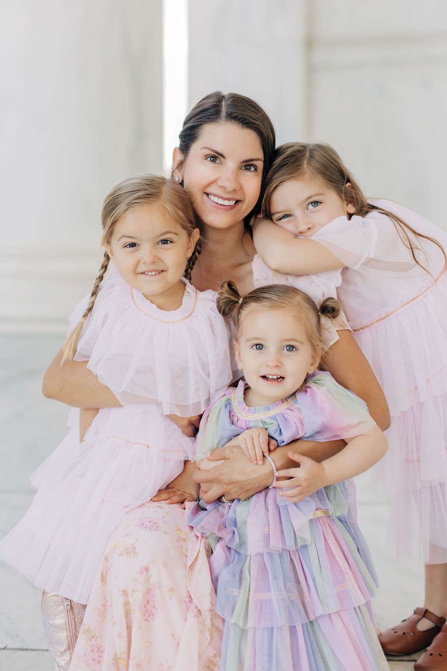 A smiling mother poses with her three young daughters in pink dresses at a white marble monument.