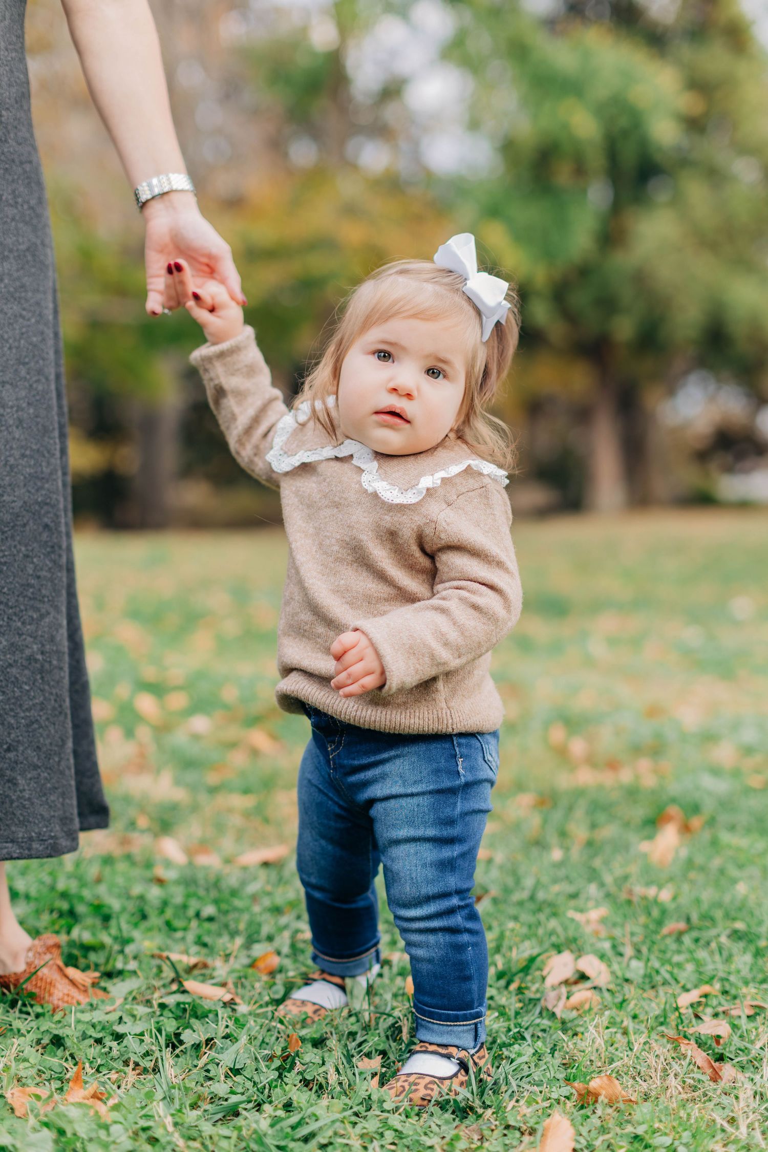 Toddler girl in beige sweater and jeans holds mother's hand while walking on grass in autumn park.