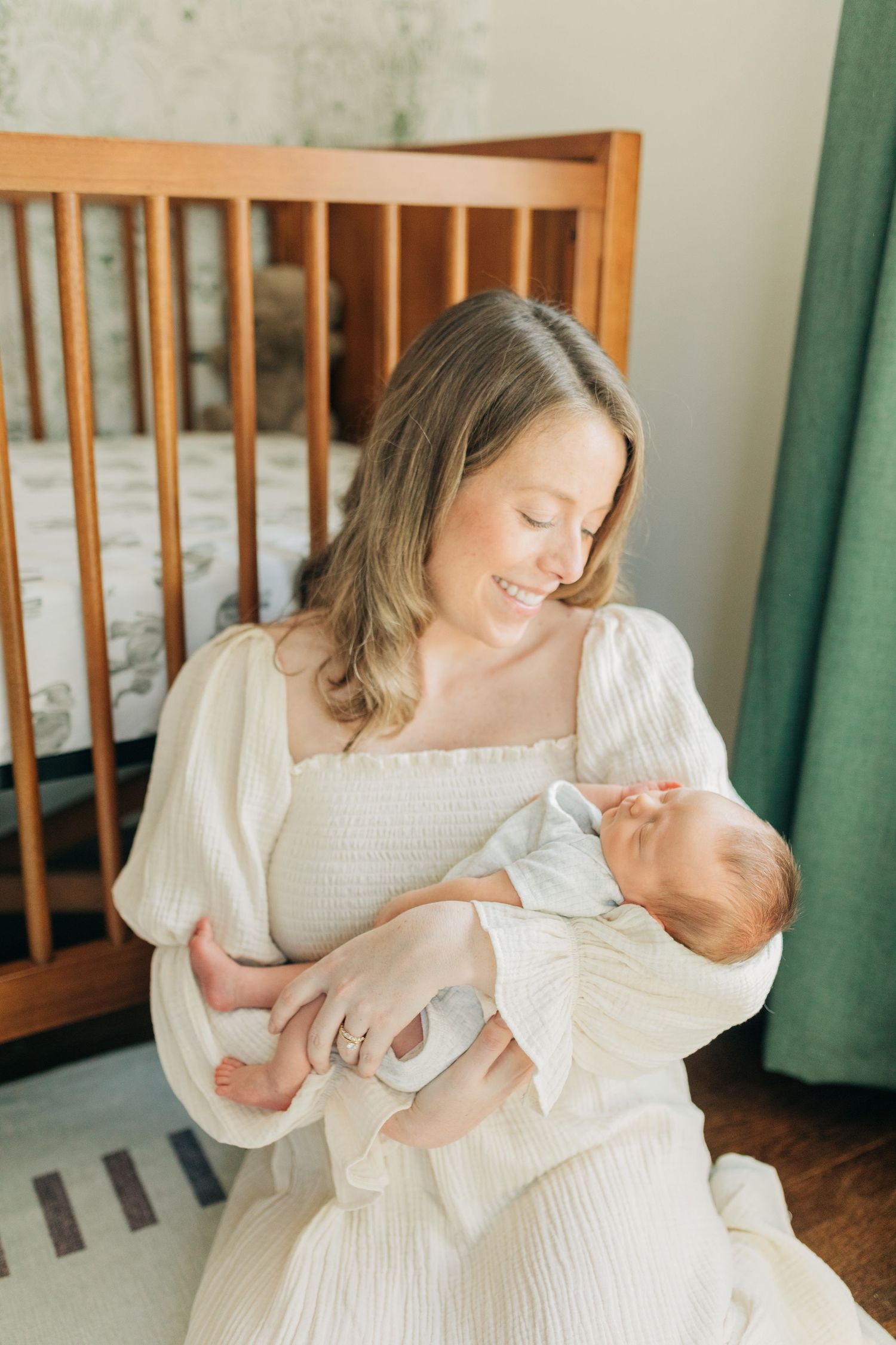 Mother in white dress holds sleeping newborn baby near wooden crib.