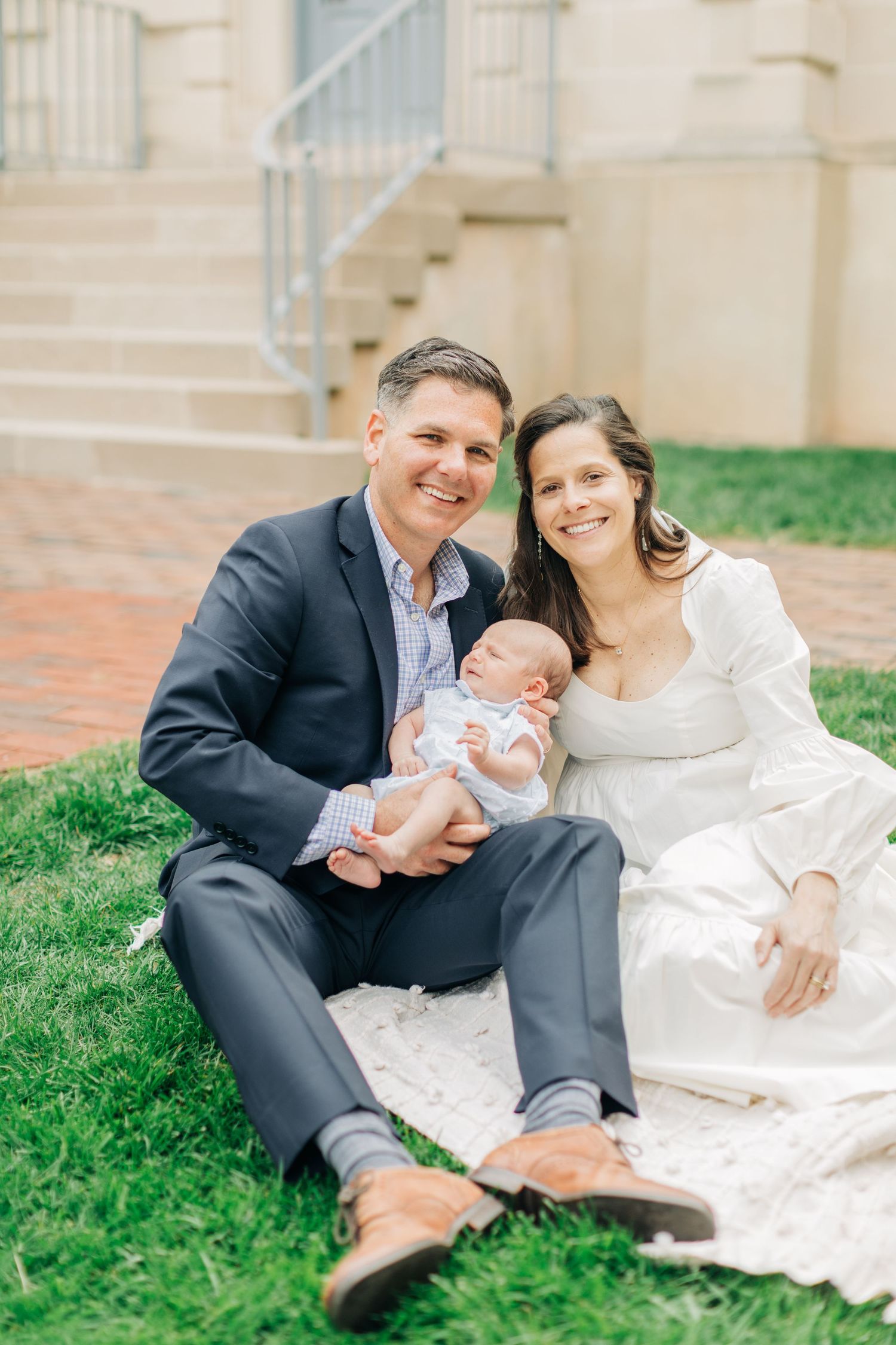 Family sits together on lawn near brick building during portrait session.