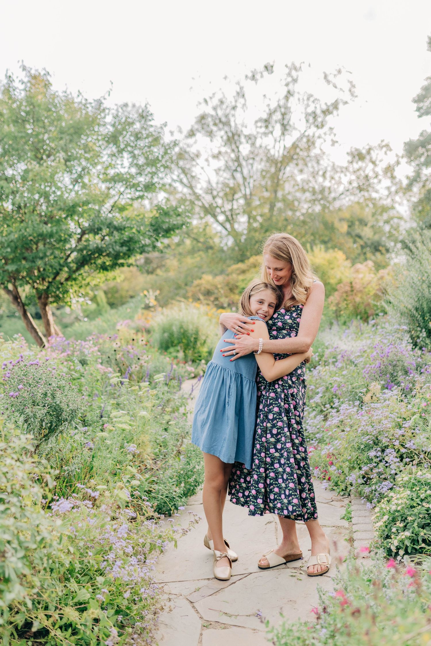 Two people share an affectionate moment while standing on a stone path in a flowering garden.