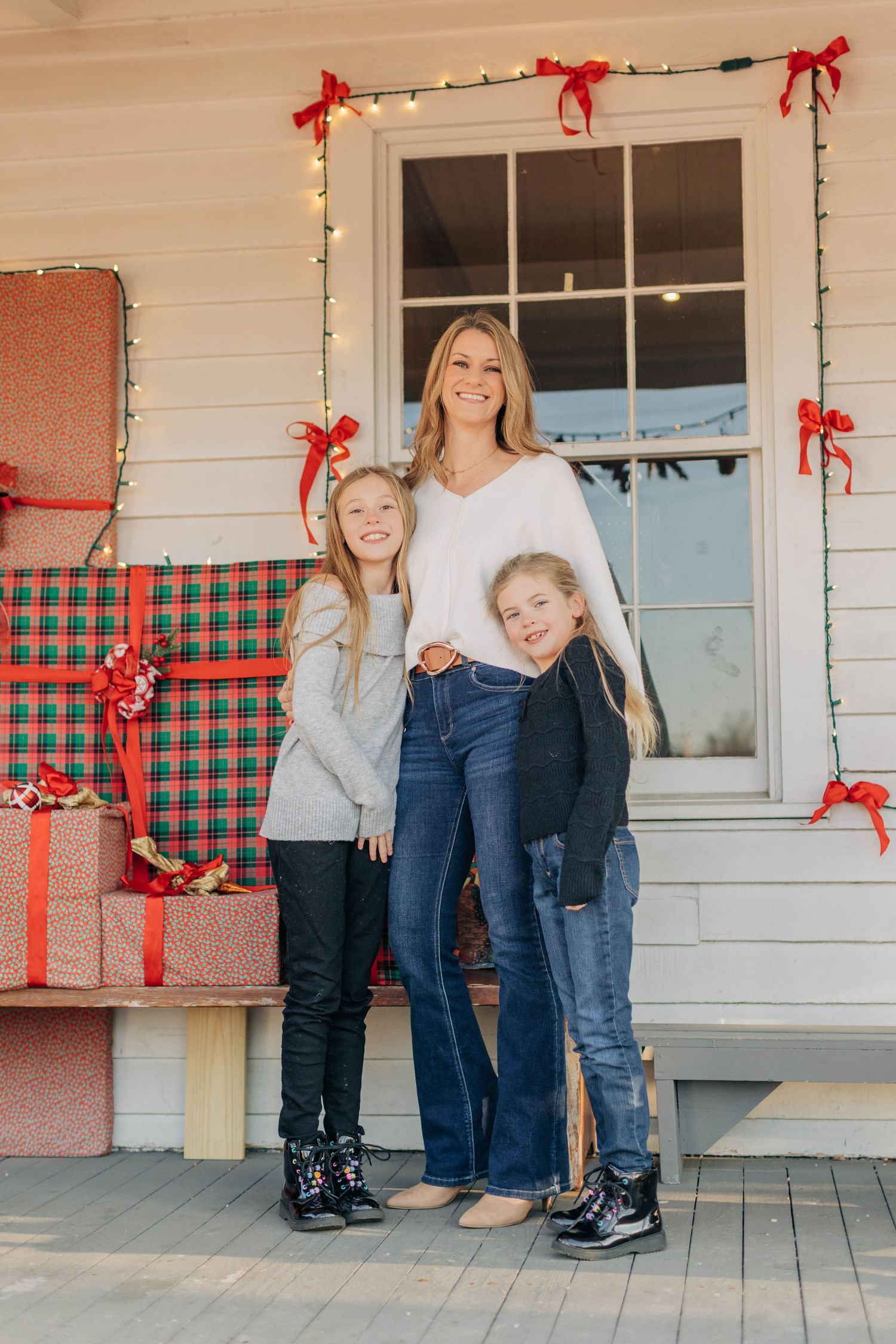 A woman and two girls pose on a Christmas-decorated porch with wrapped gifts and holiday lights.