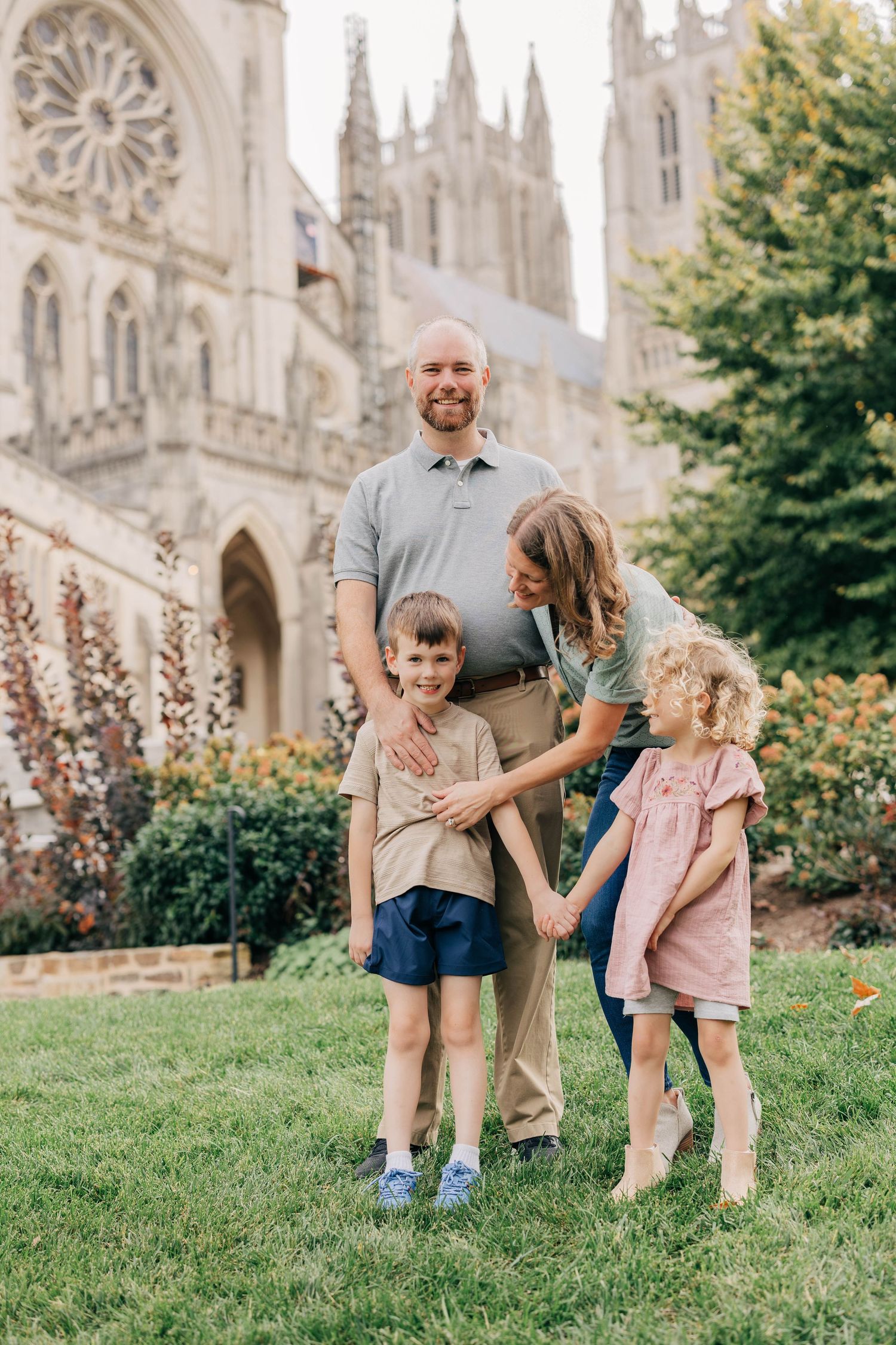 Smiling family of four standing together on a green lawn in front of a Gothic stone cathedral with tall spires on a sunny day.