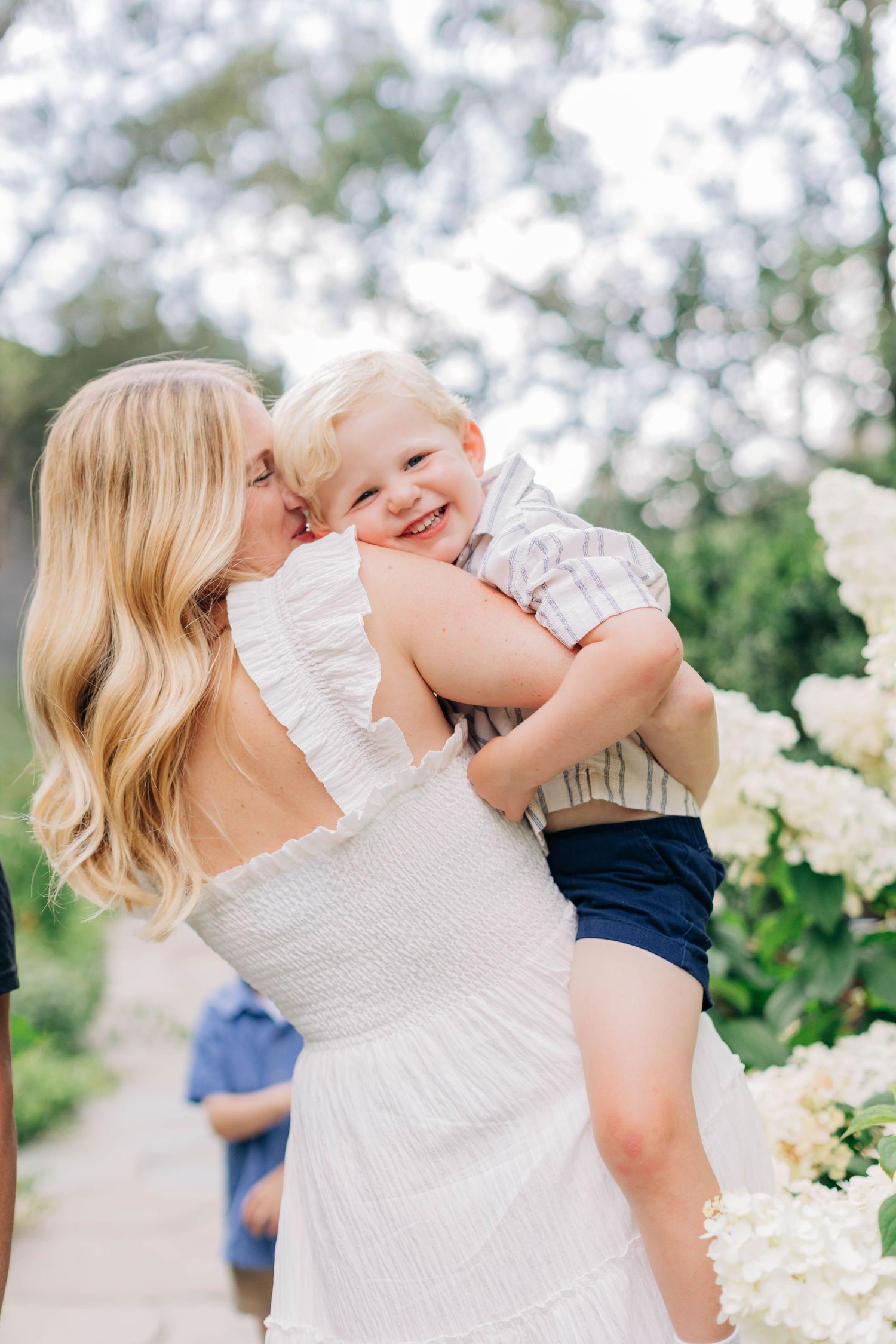 Blonde mother in white dress kissing smiling toddler boy outdoors near white hydrangeas.