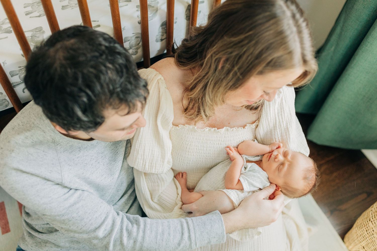 Family bonding moment captured in soft natural light by wooden crib and green curtains.
