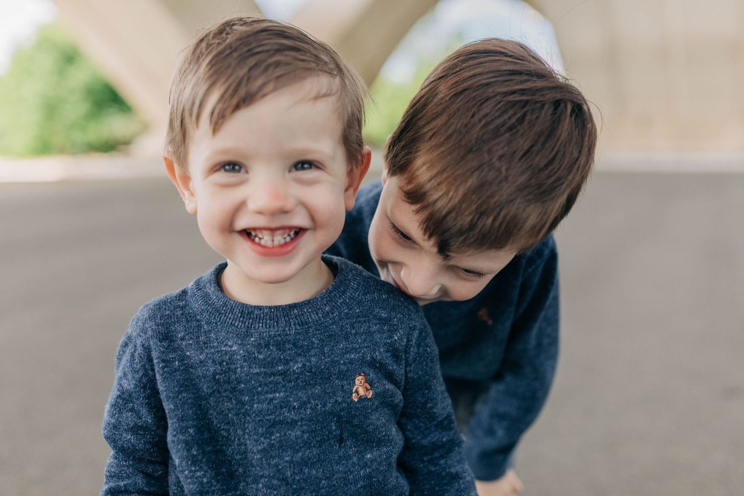 Two children in matching navy sweaters sharing playful moment under concrete structure.