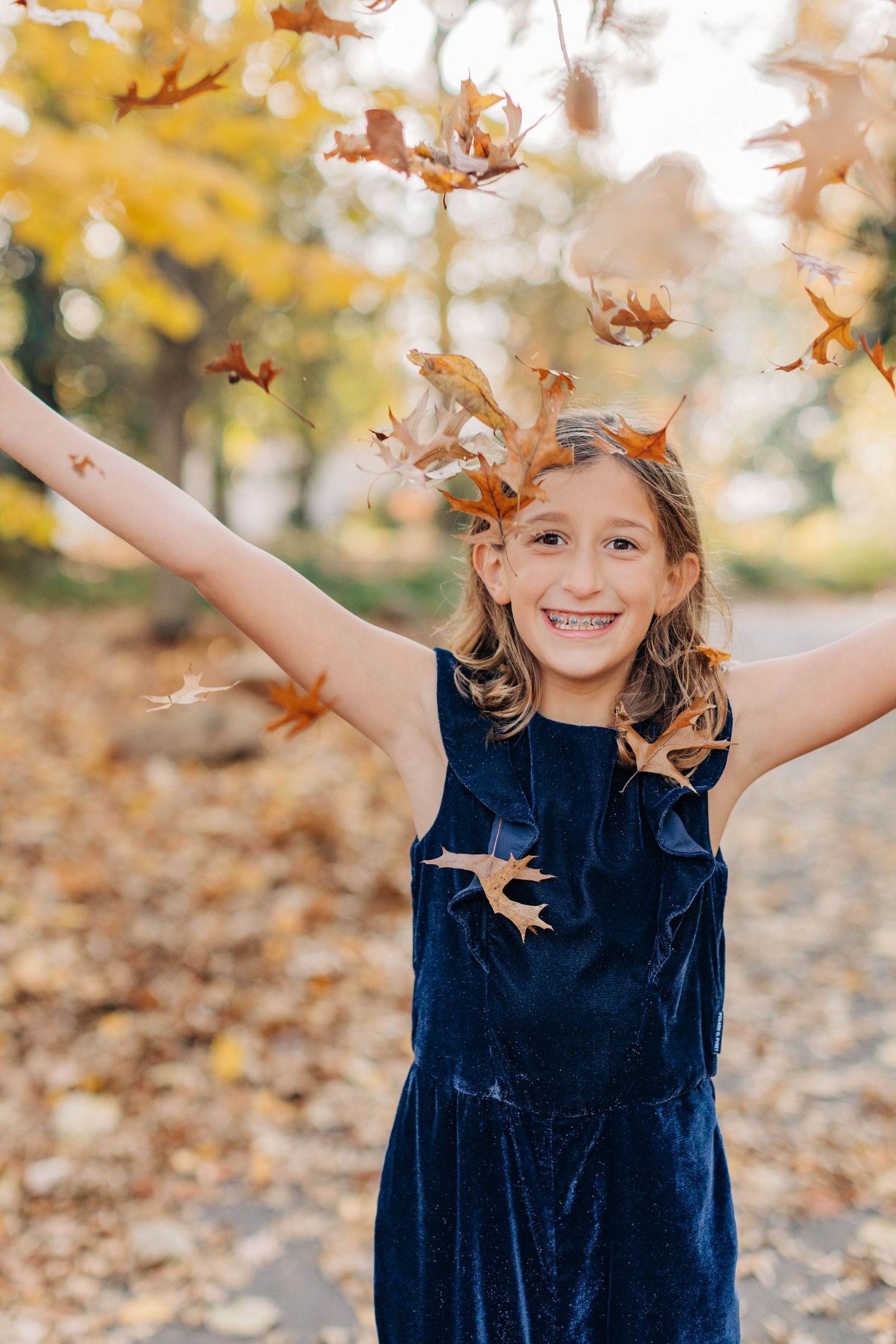 A child in a navy dress plays with falling autumn leaves in a park.