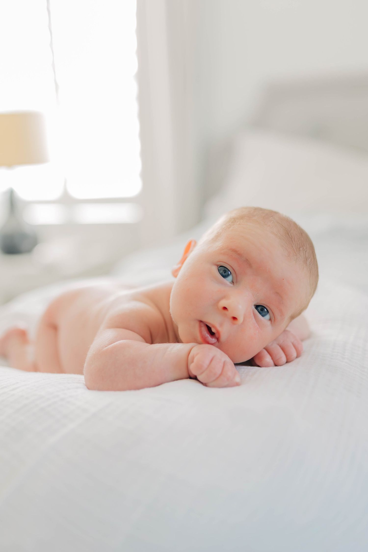 A baby lies on white bedding while looking up with bright blue eyes.