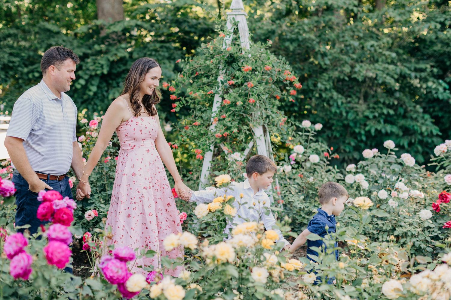 Family walking through a colorful rose garden, parents holding hands while two young boys explore the blooming flowers.