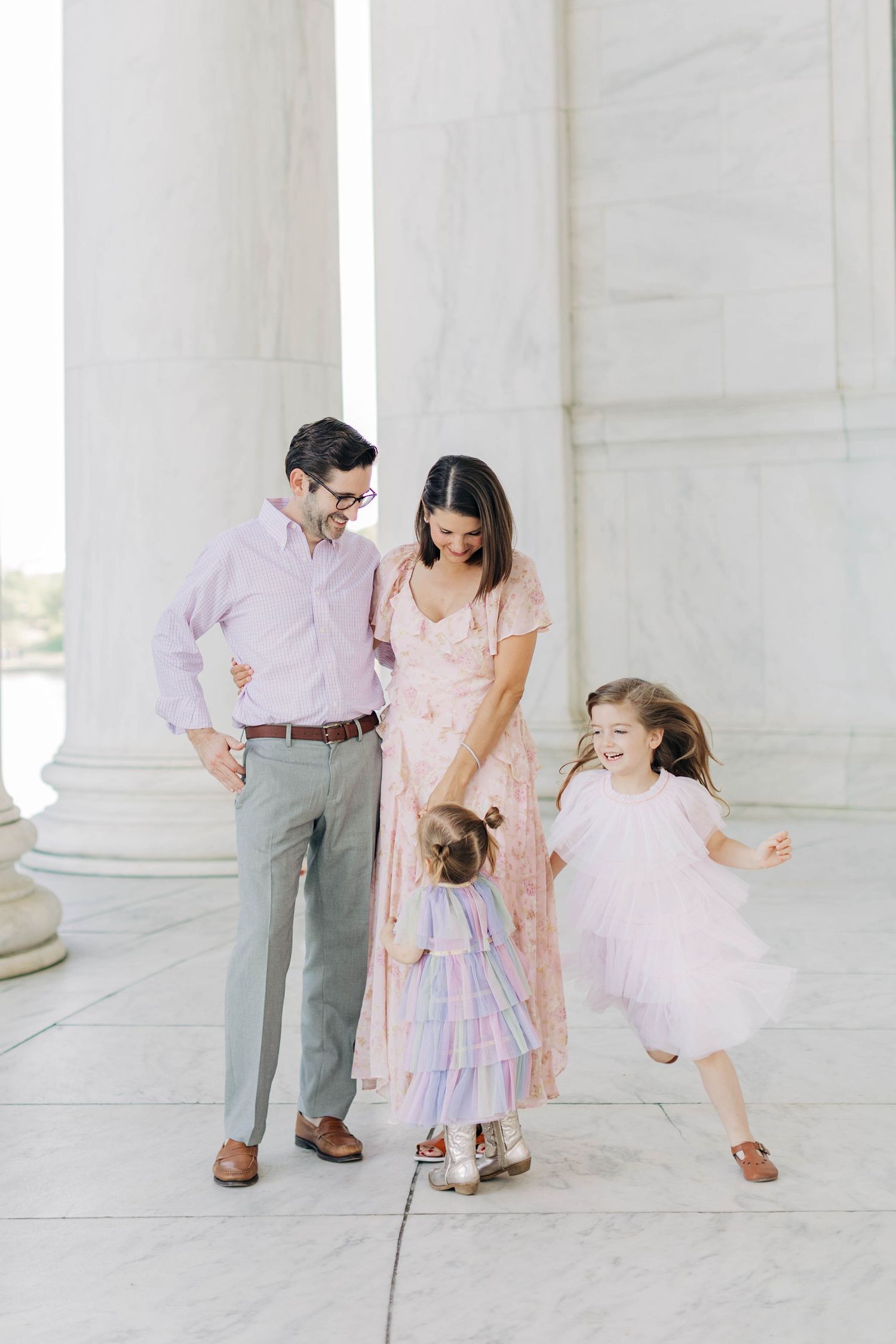 Family of four posing at Jefferson Memorial, parents watching two young girls in pastel dresses laugh and play.