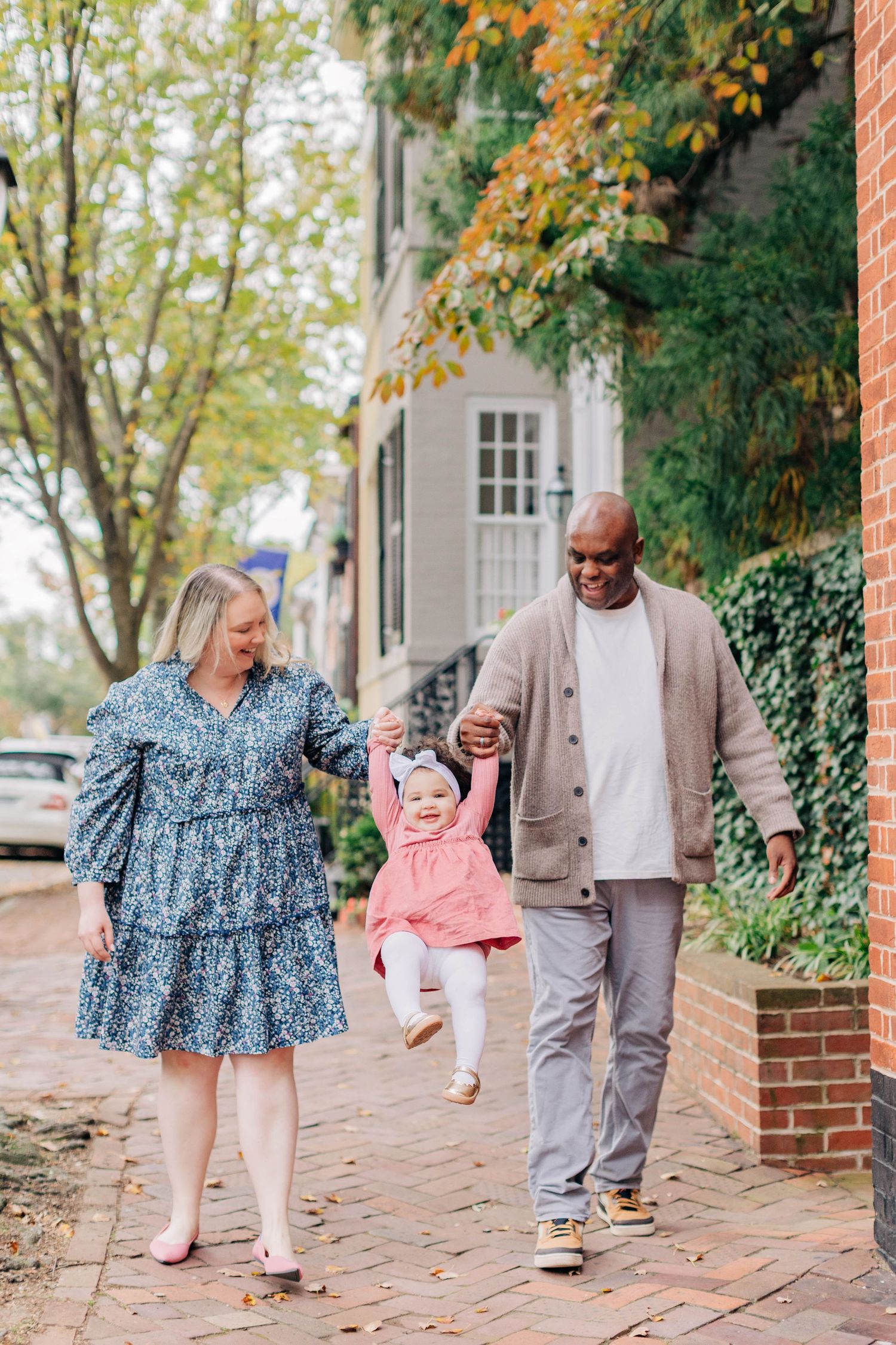 A joyful interracial family swings their toddler girl between them while walking on a brick sidewalk in autumn.
