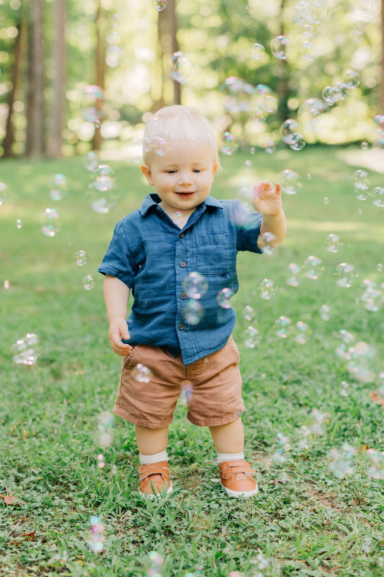 Toddler boy in denim shirt and brown shorts playing with bubbles outdoors on green grass in a sunny park.
