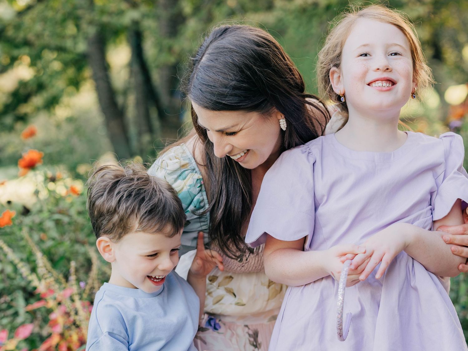 A mother smiles warmly with her two young children in a colorful garden, wearing pastel outfits.