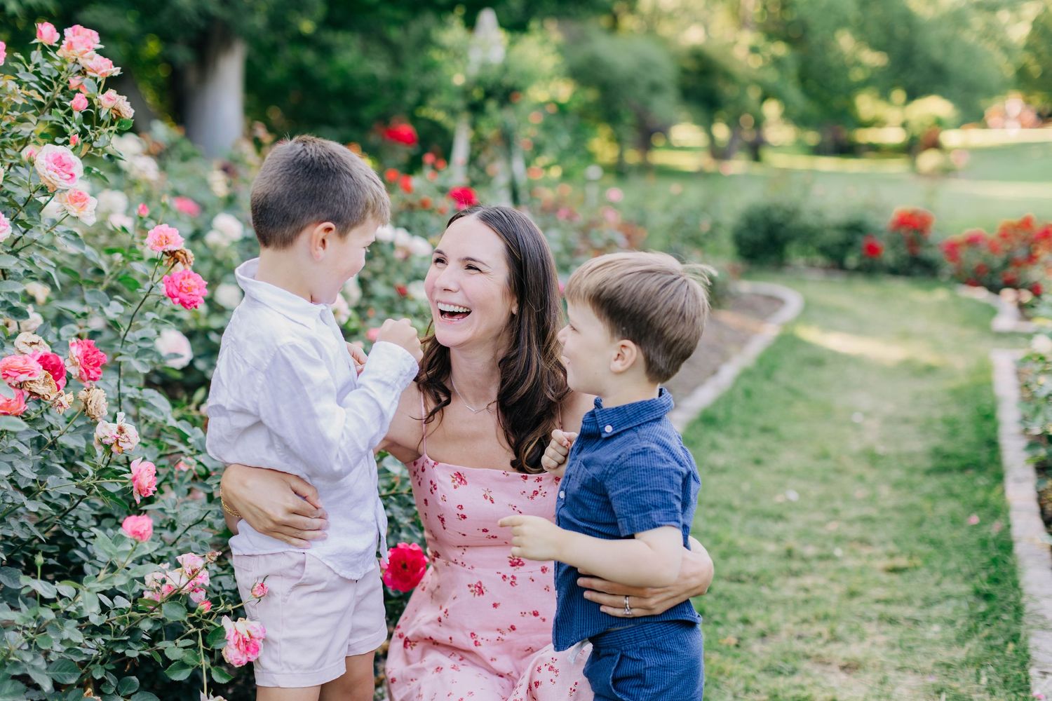 A laughing mother in a pink floral dress holds her toddler son while her older boy hugs her in a rose garden.