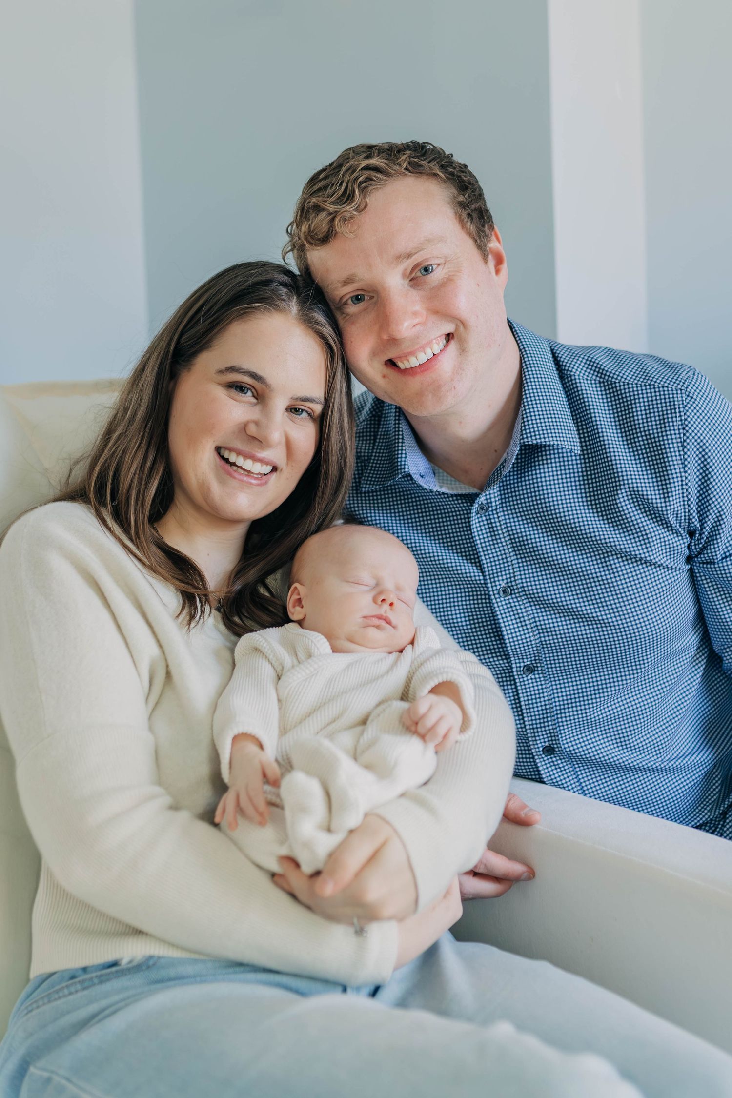 Smiling couple sitting in chair holding sleeping newborn baby in white outfit.
