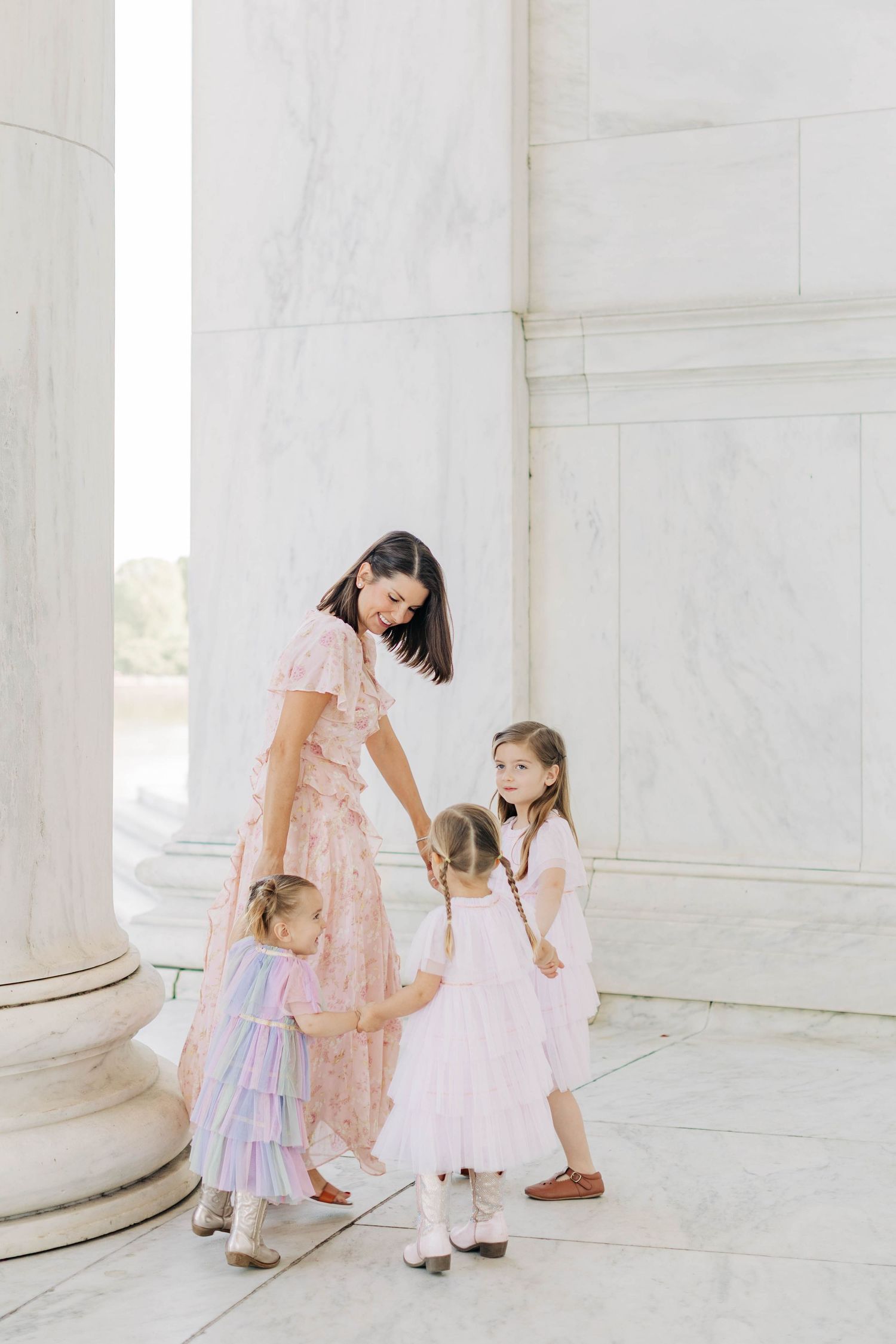 A mother in a pink floral dress interacts with her two young daughters in matching pink dresses near marble columns.