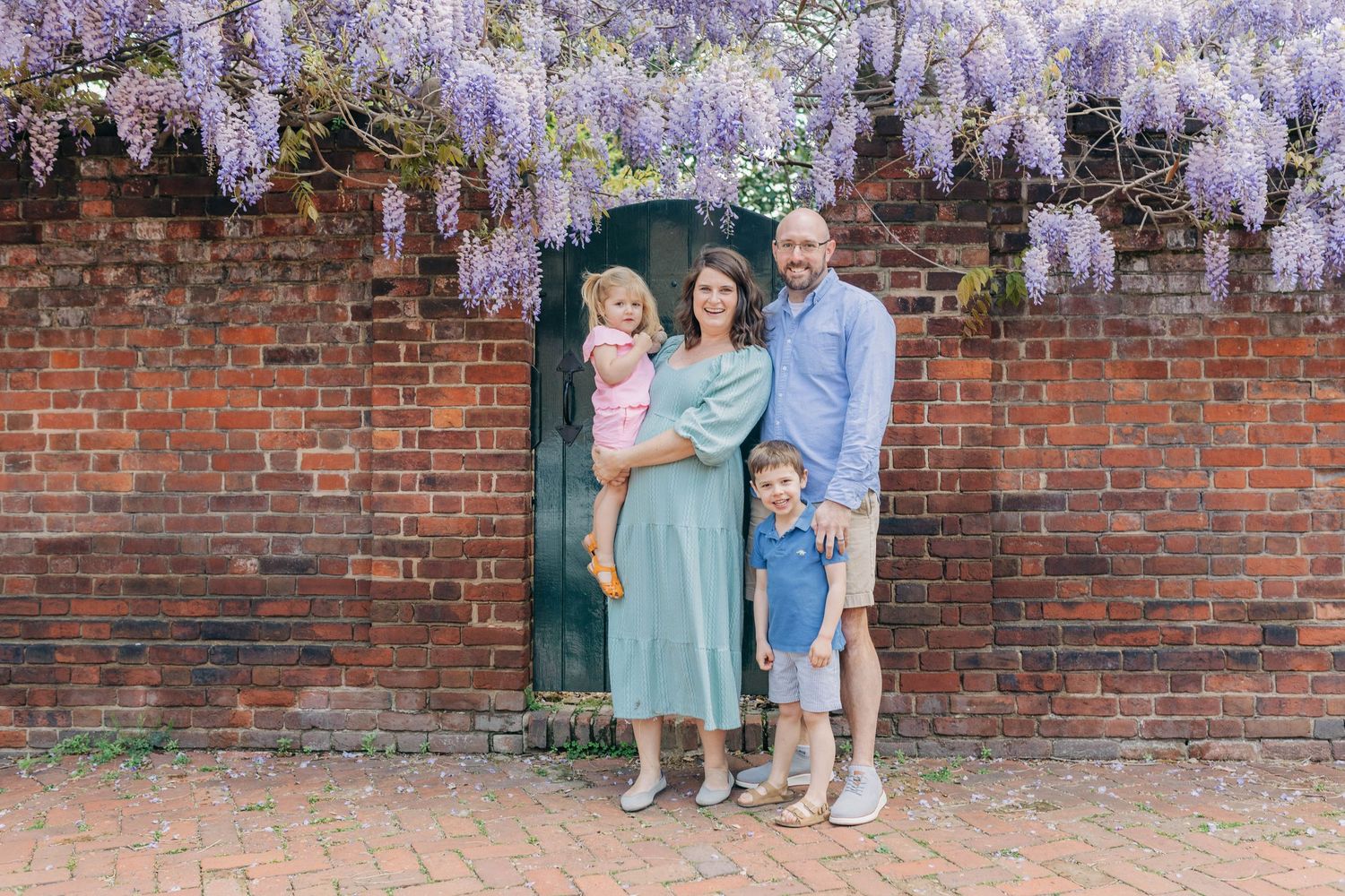 Family of four posing under wisteria blooms by a brick wall with a green door in a garden setting.