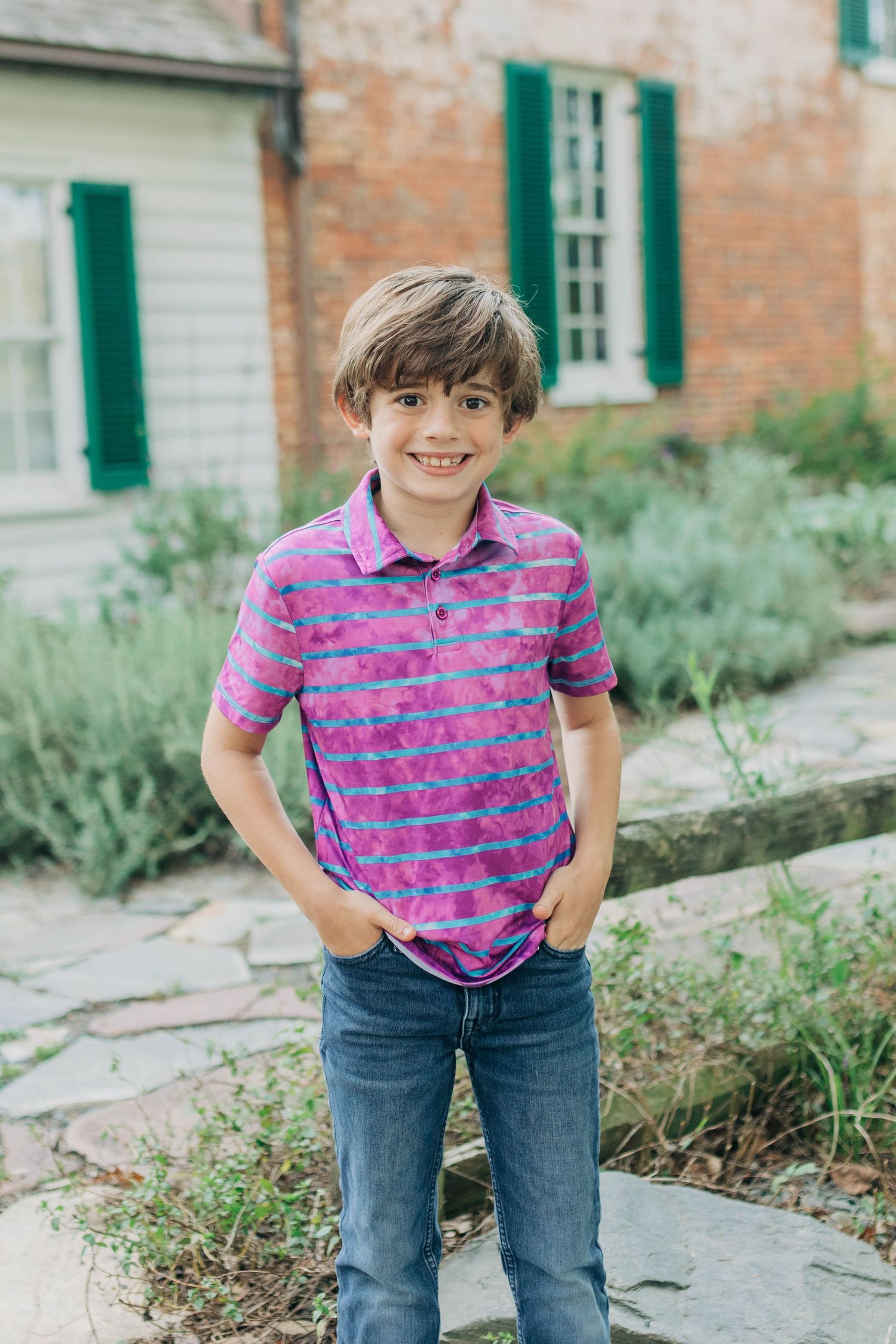 Child in pink and blue striped polo shirt smiling while standing near historic building.