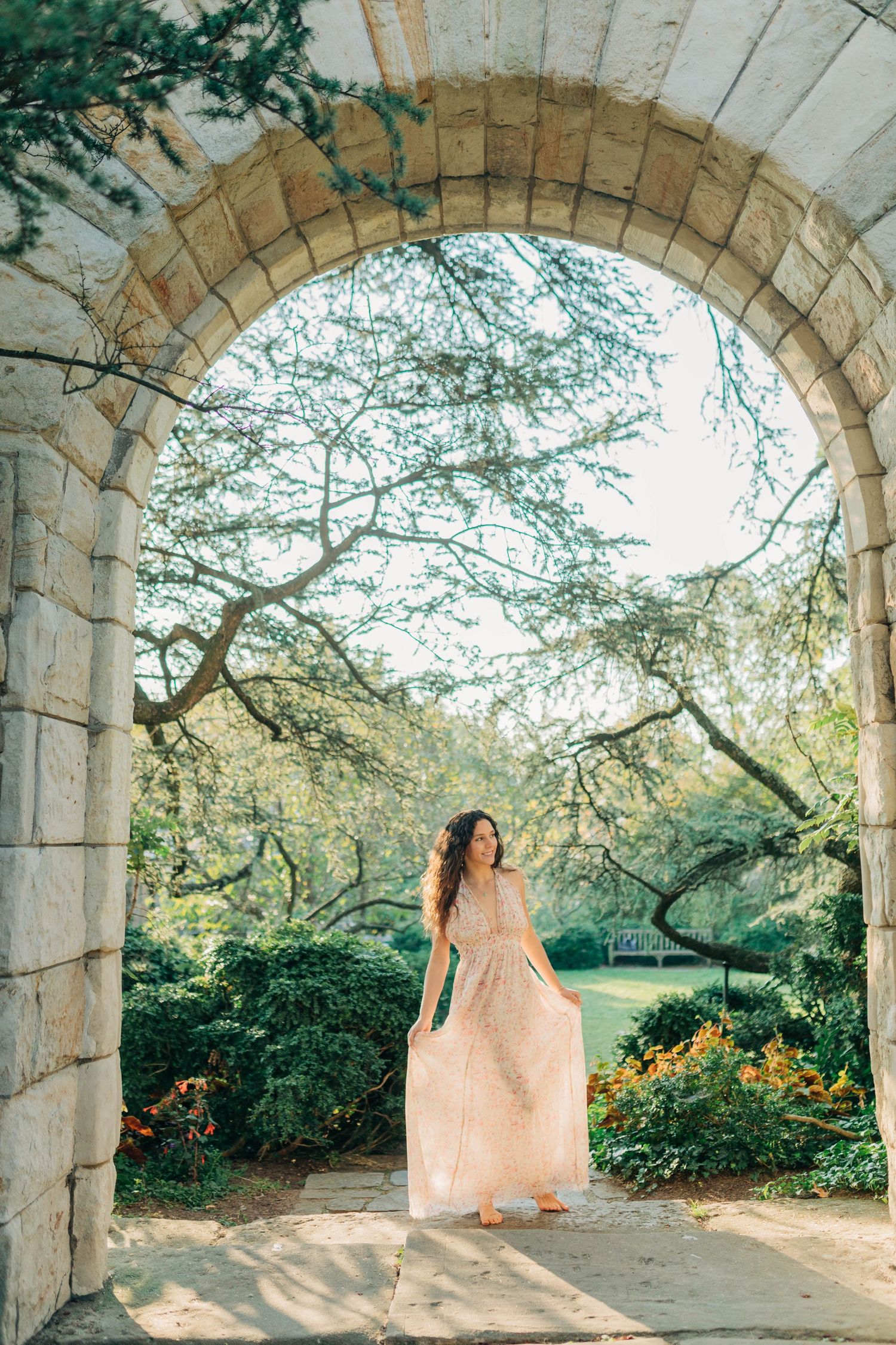 A flowing pink dress catches sunlight under stone arches in a garden setting.