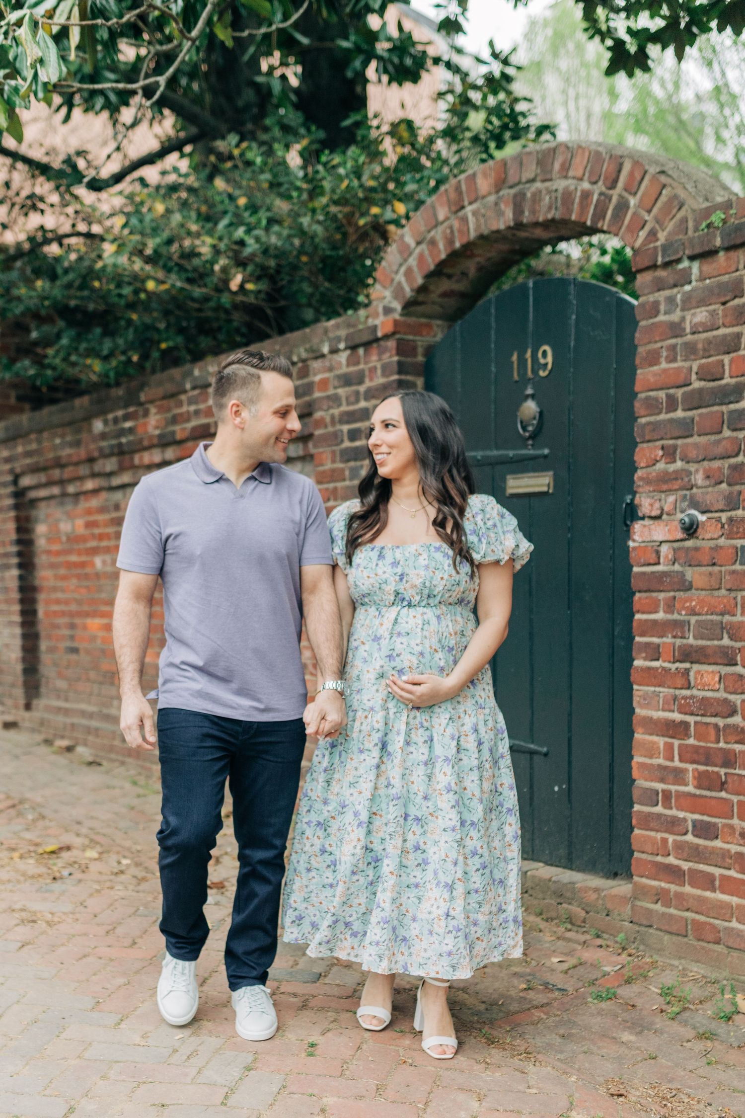 A couple walks and poses playfully along a brick wall with dark green doors.