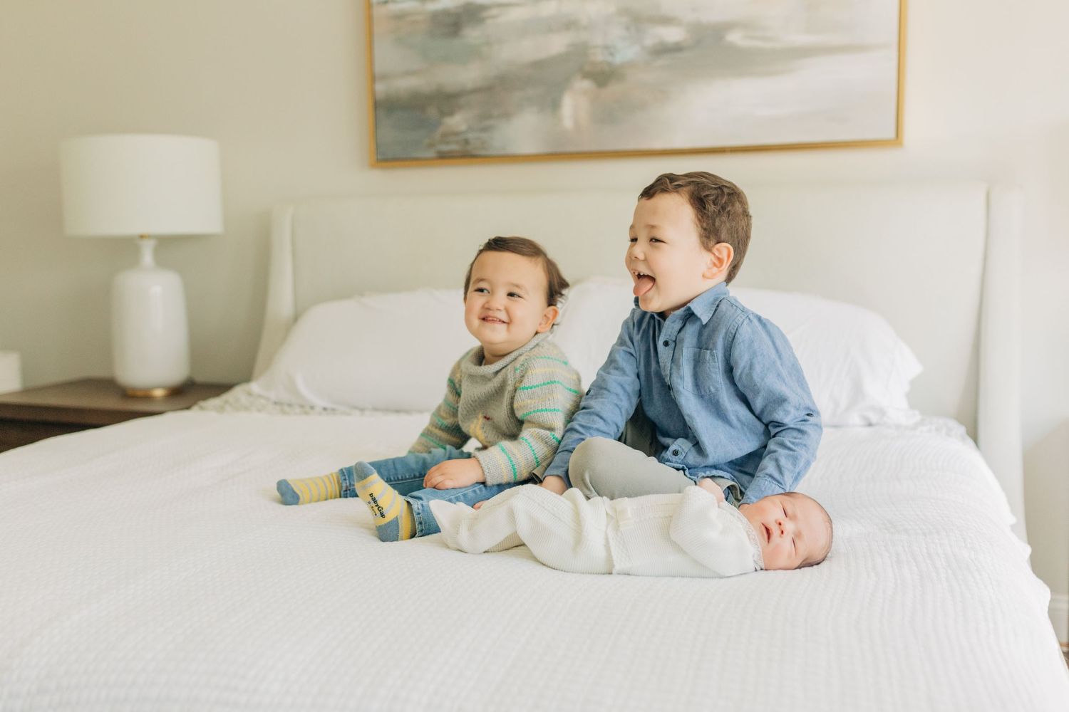 Three children lay together on a white bed with a serene abstract artwork hanging above.