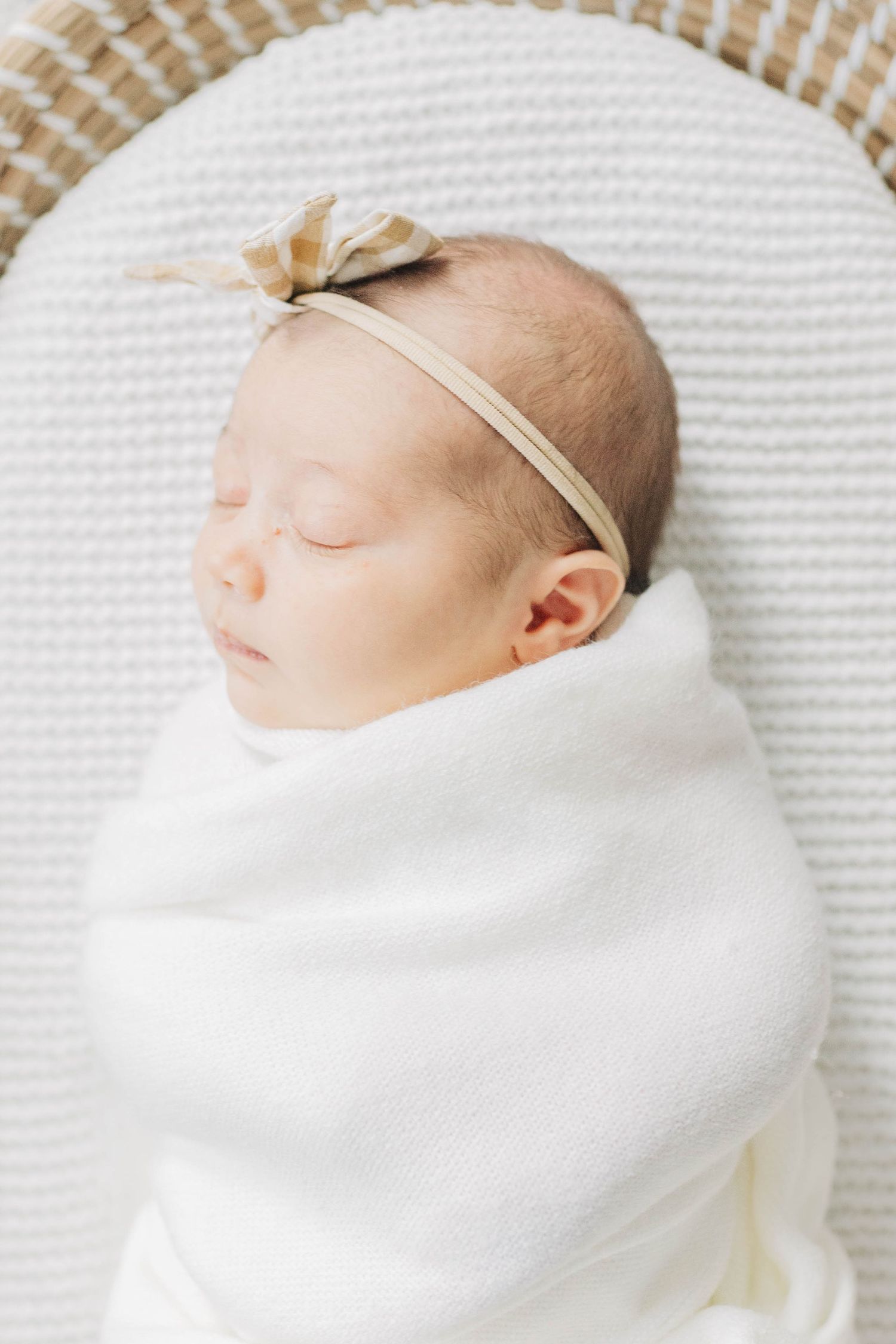 Sleeping newborn swaddled in white blanket wearing delicate beige bow headband on textured surface.