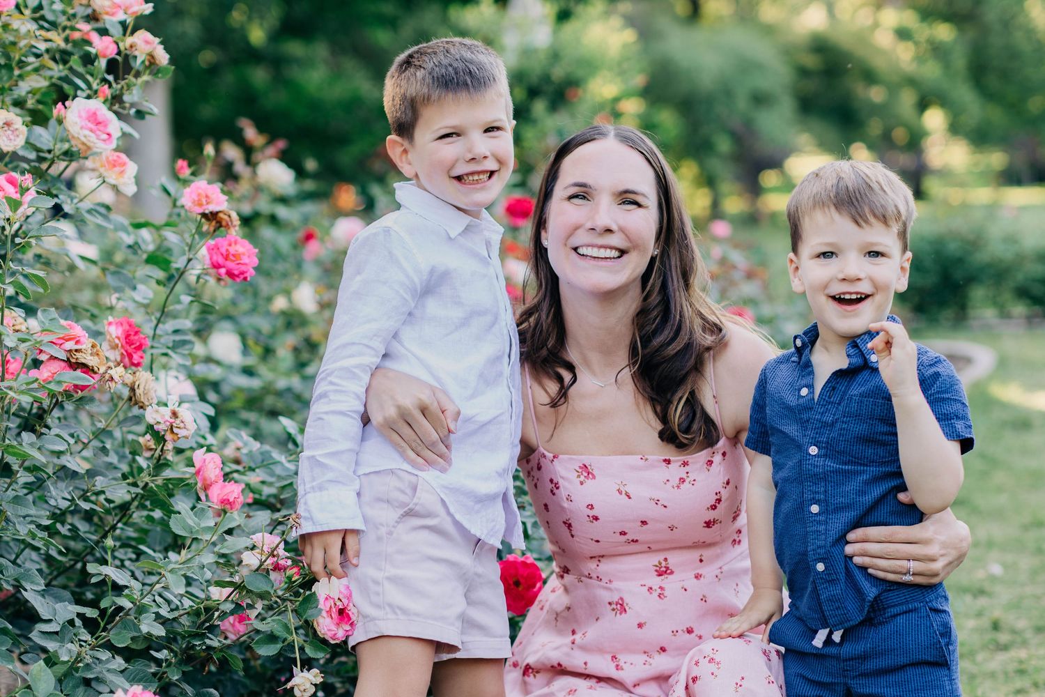 A happy mother in a pink dress poses with her two young sons beside colorful roses outdoors.