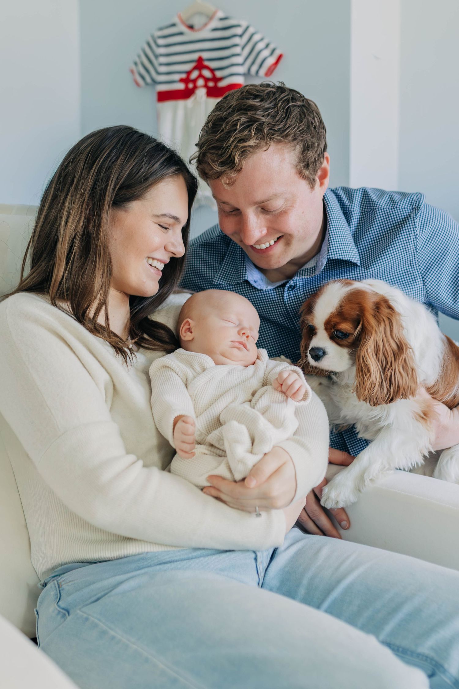 Smiling parents sitting on sofa holding newborn baby with Cavalier King Charles Spaniel.