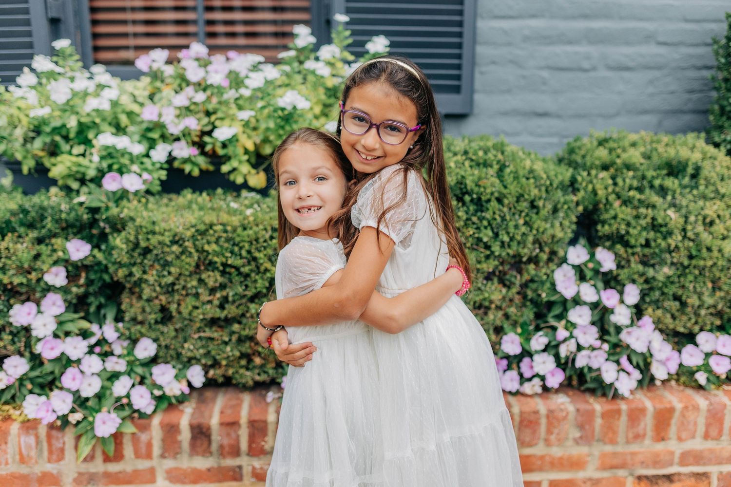 Two young girls in white dresses hugging and smiling in front of blooming pink hydrangeas on a sunny day.