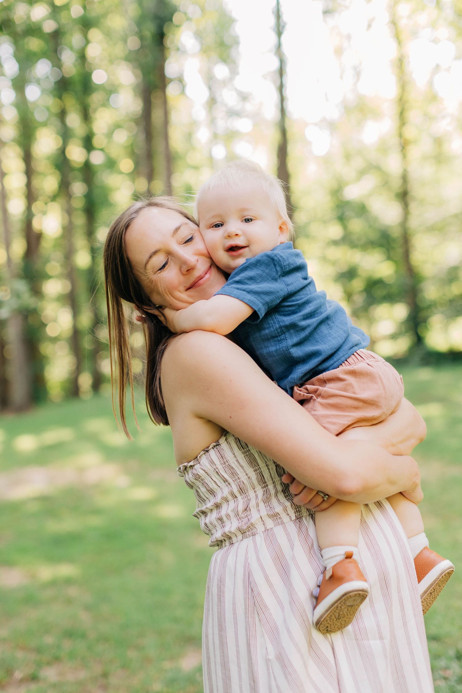 A smiling mother tenderly hugs her happy baby boy outdoors in a sunny green park setting.
