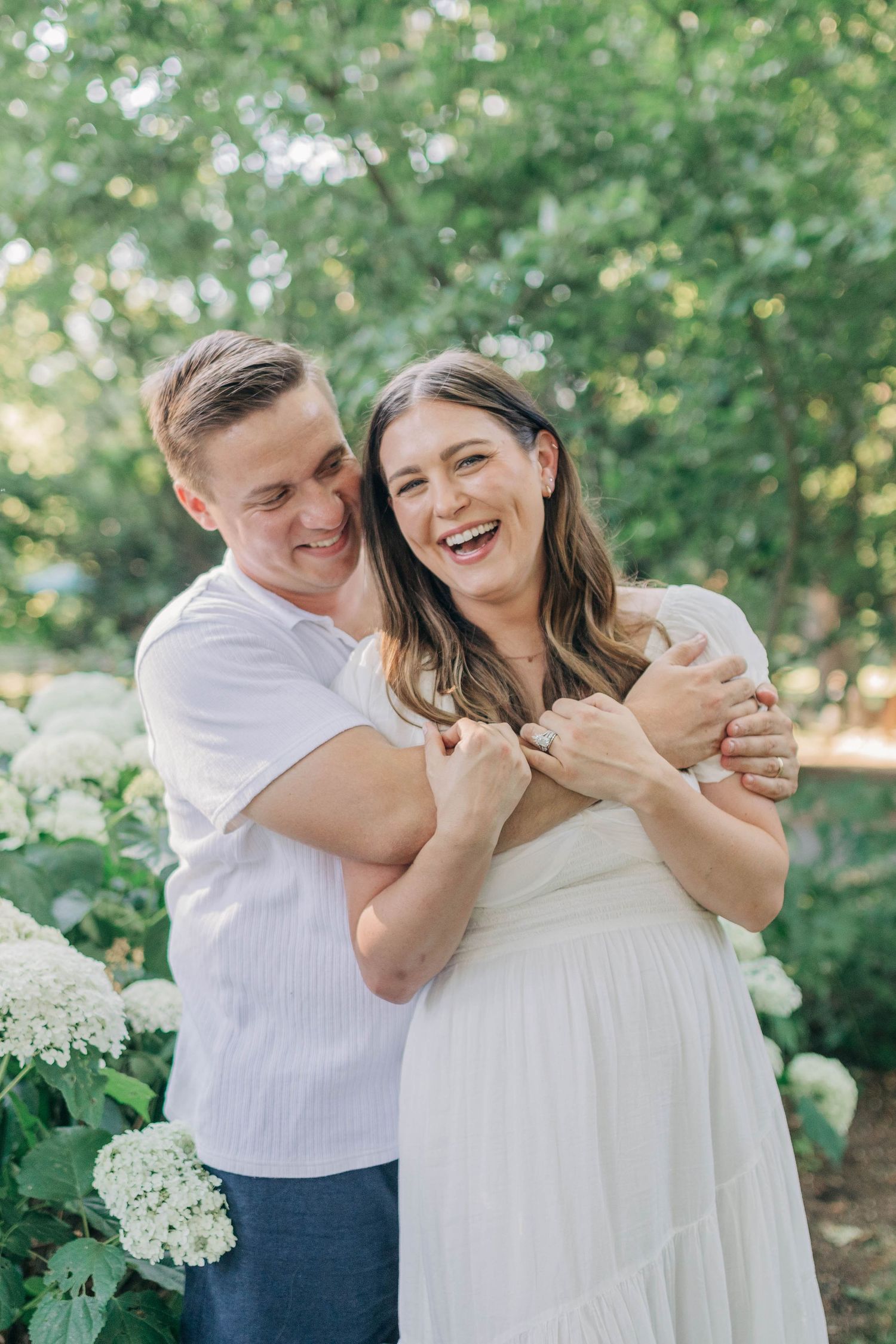 Family shares intimate laughing moments together in white outfits against flowering garden backdrop.