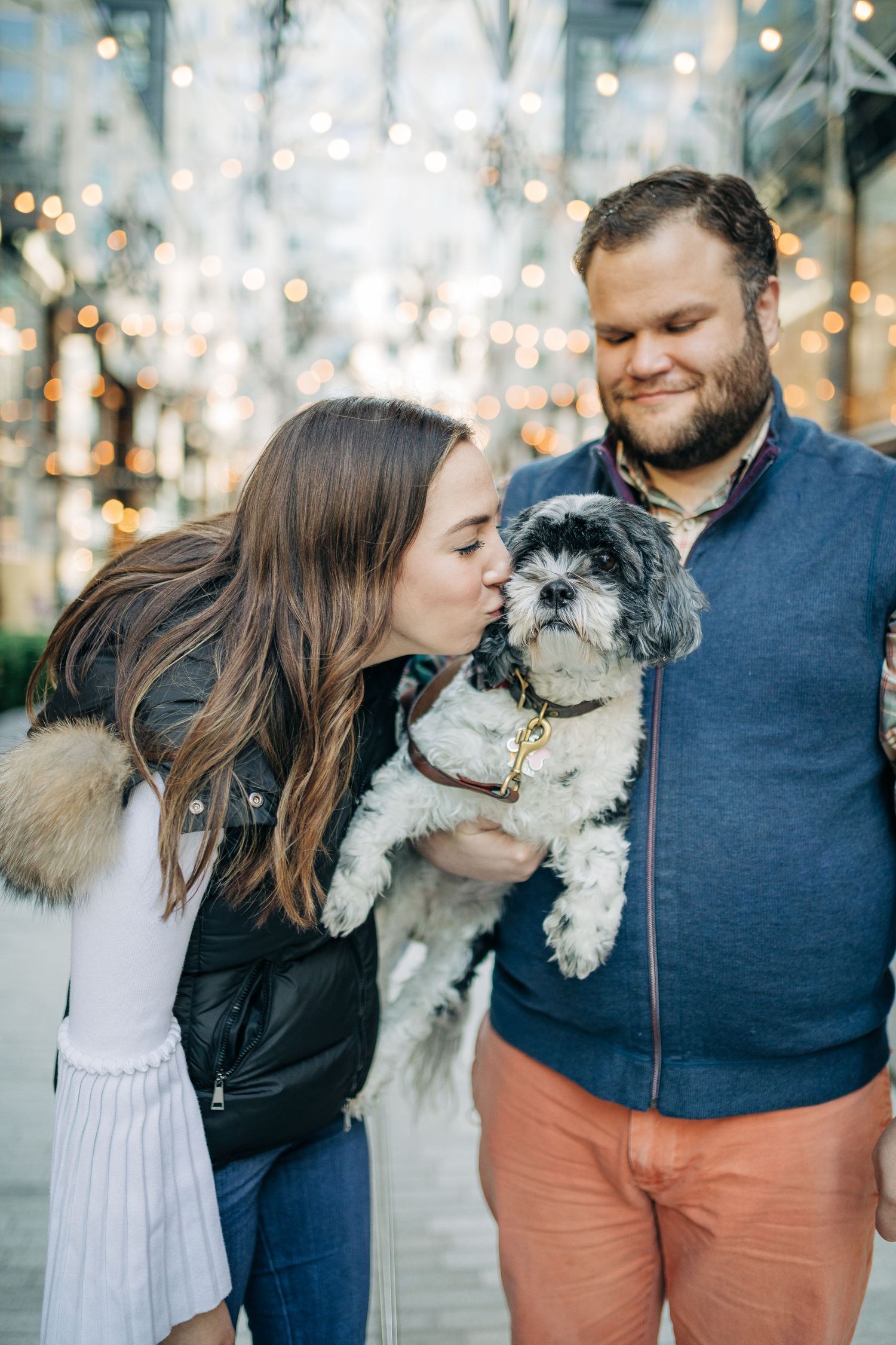 A couple poses with their small white and gray dog under festive holiday lighting.