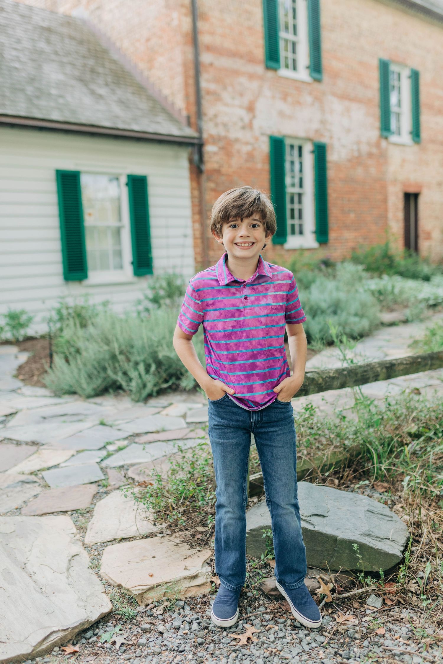 Child in purple striped shirt posing along stone pathway near historic brick building with green shutters.