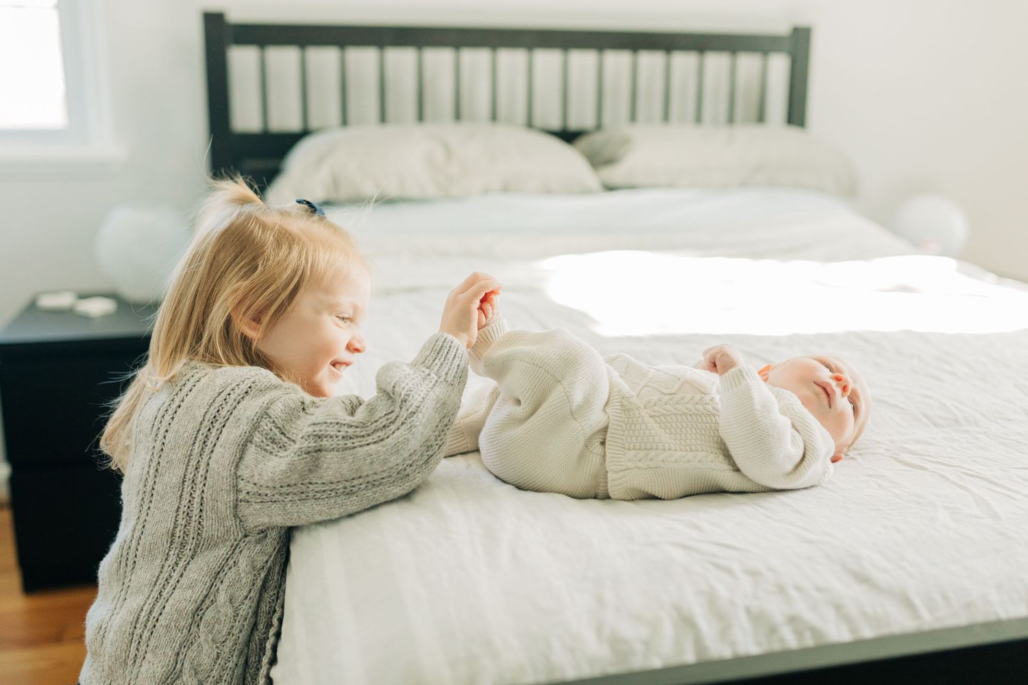 A child in a gray sweater plays with a baby on white bedding.