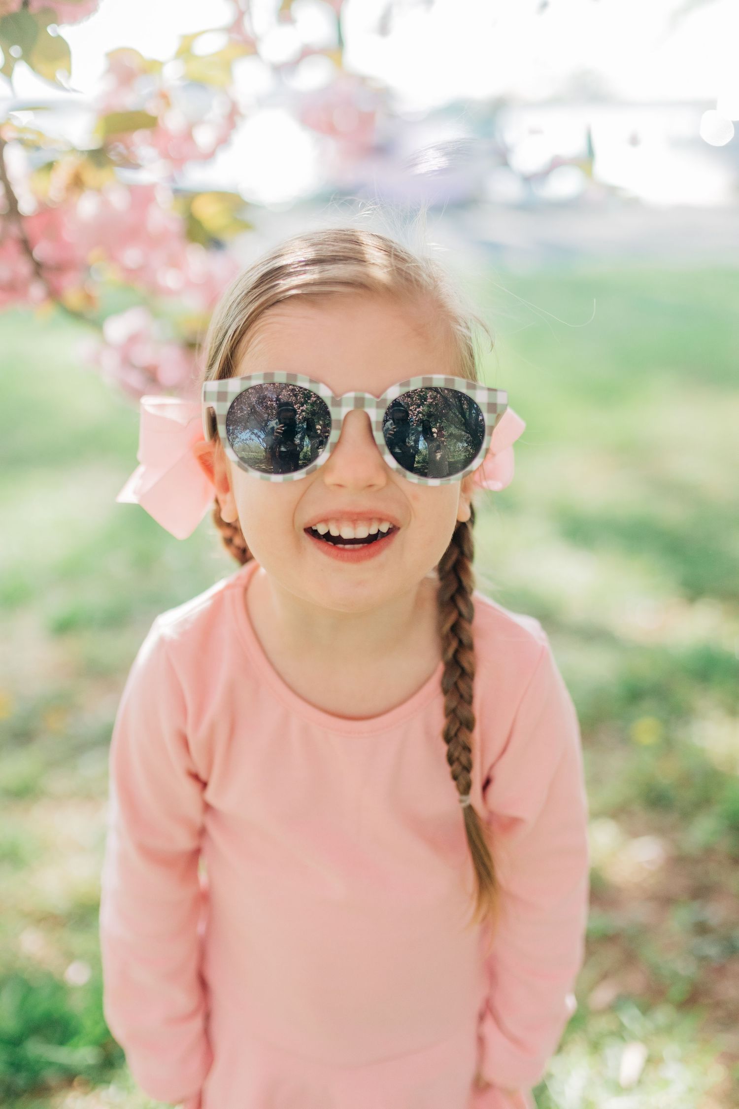 Person in pink sweater and sunglasses smiles brightly under spring blossoms.