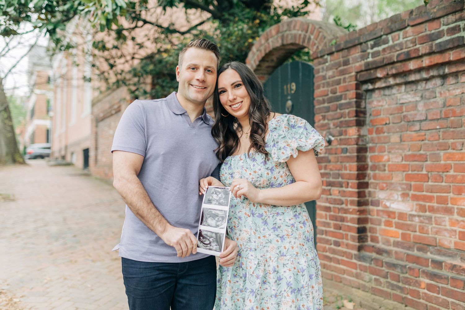 A couple shares loving moments while holding ultrasound photos on a brick-lined path.