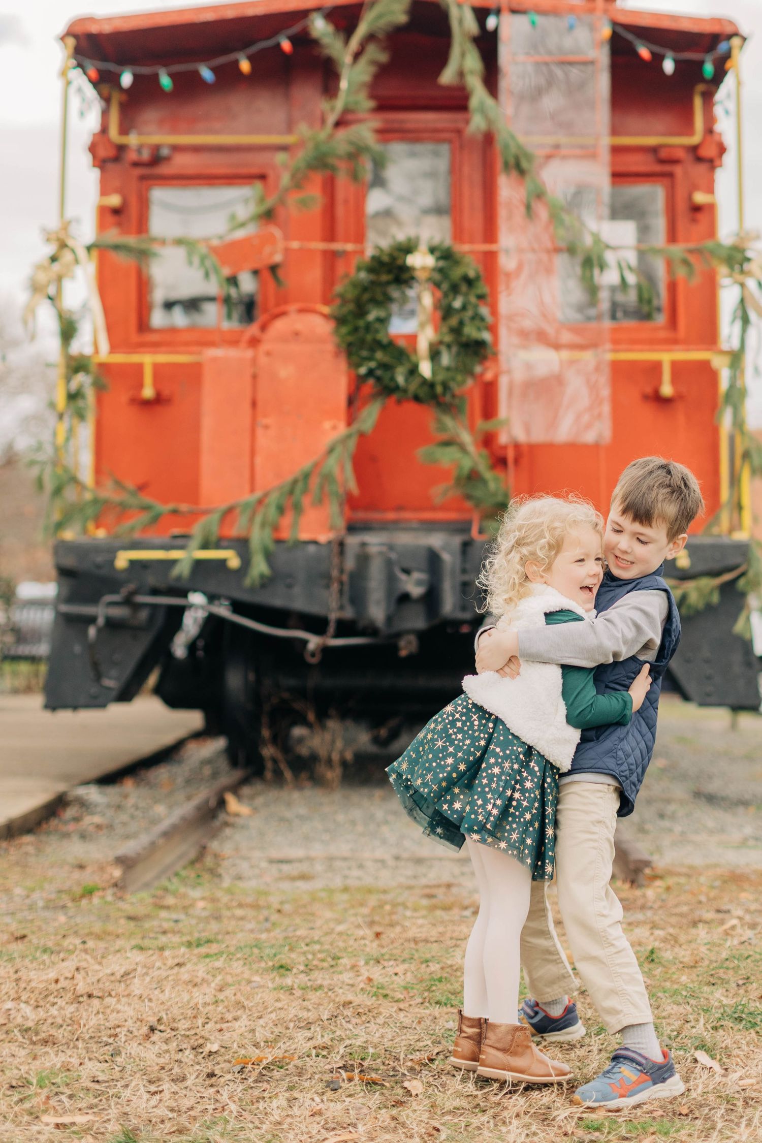 Two young children hugging in front of a red Christmas-decorated train car with a holiday wreath.