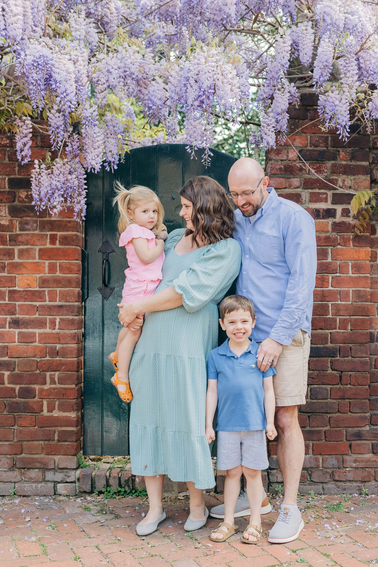 Happy family of four poses by a brick wall green door under blooming purple wisteria flowers.