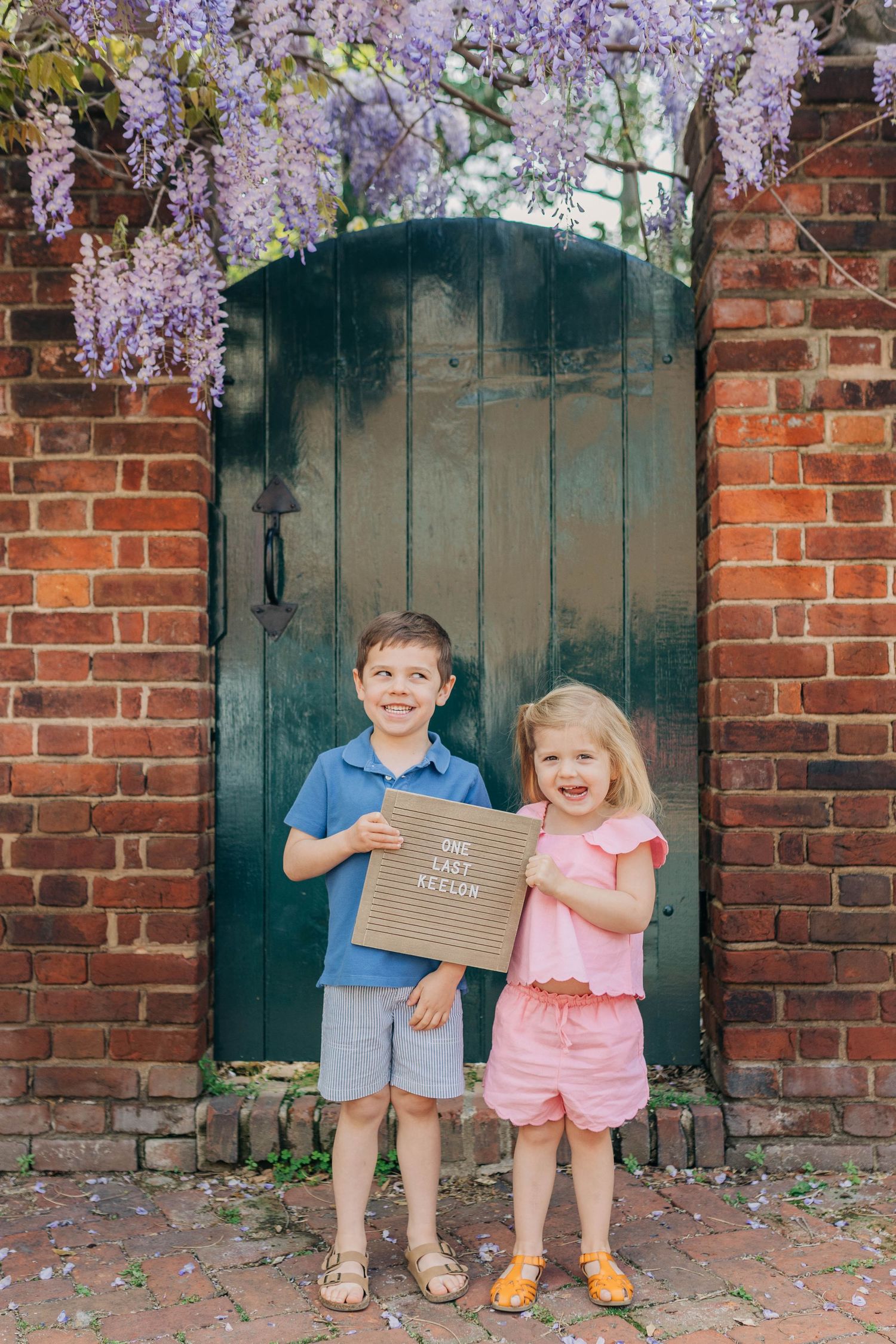 Two smiling children hold a letter board sign in front of a green door with purple wisteria blooms.