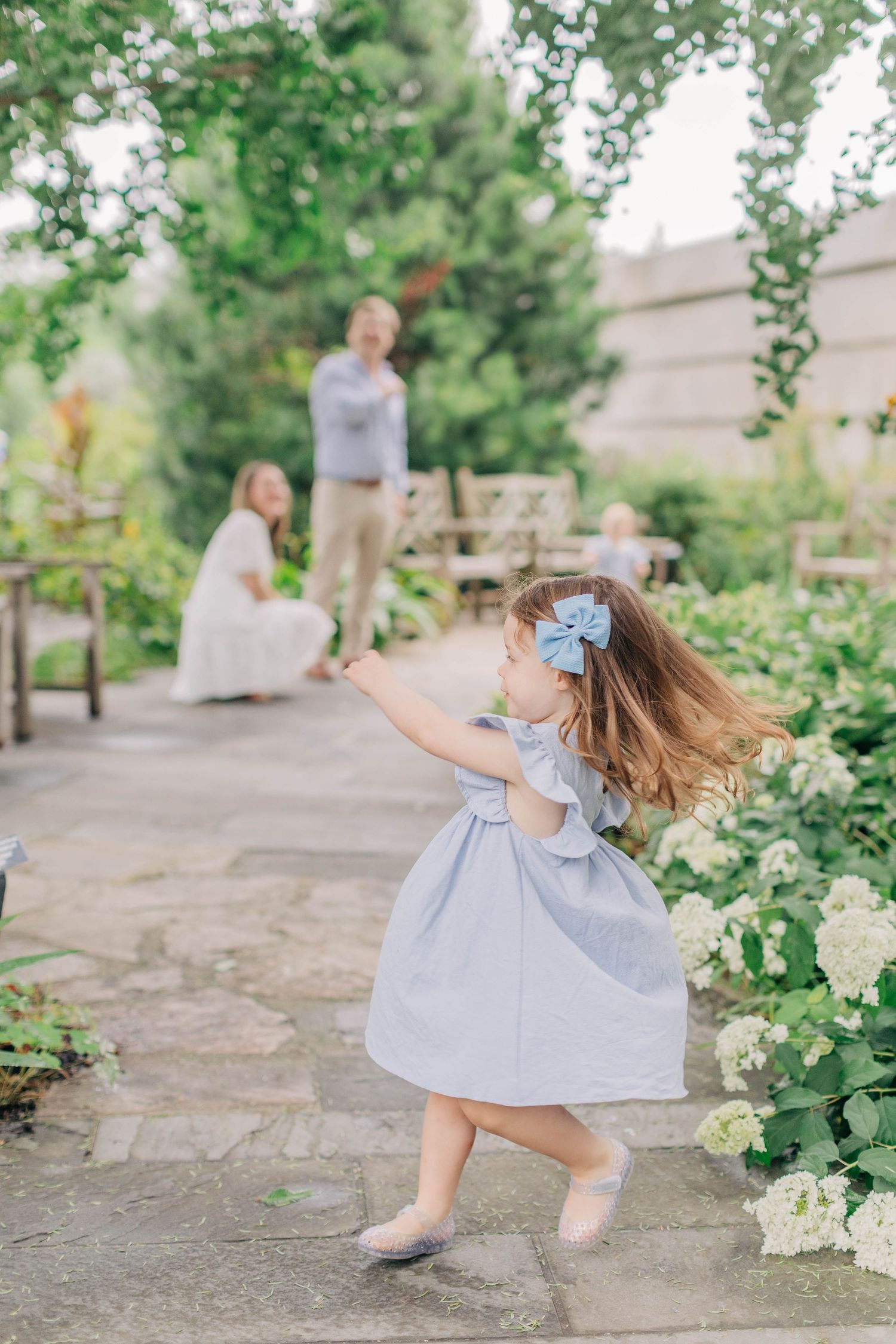A young girl in a blue dress and bow twirls on a garden path while her family enjoys the lush surroundings.
