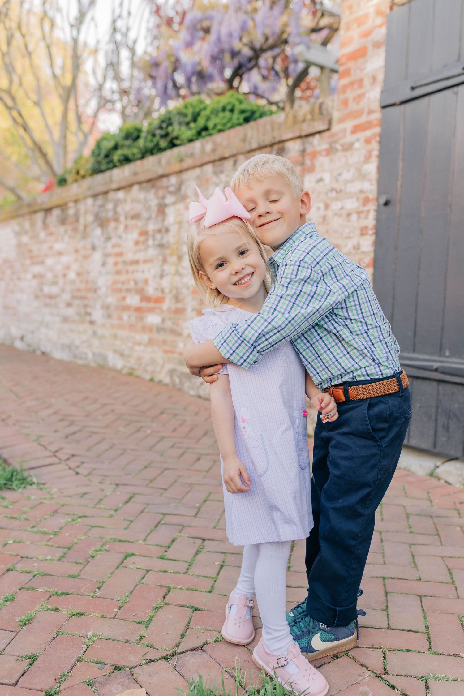 Two young blonde siblings share a sweet hug on a brick path beside a rustic wall with purple wisteria.