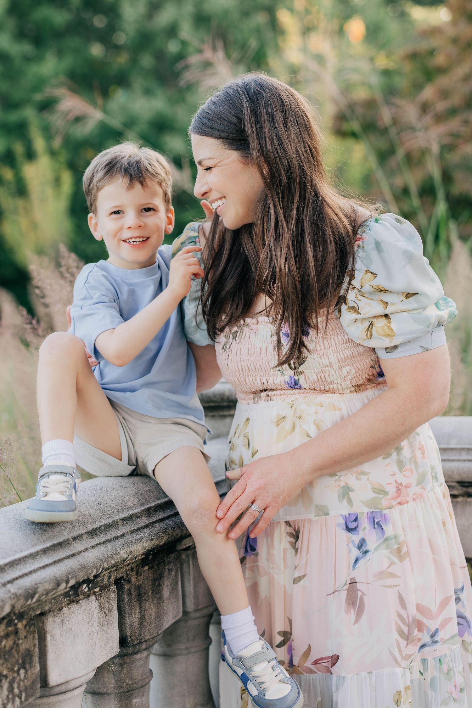 Pregnant woman in floral dress smiling at young boy sitting on stone railing outdoors in garden.