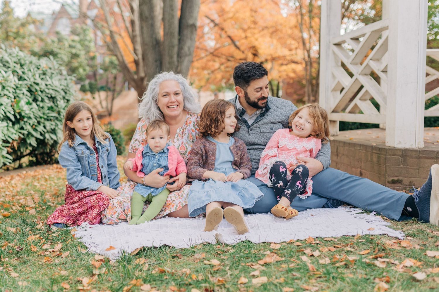 Family of six sitting on a blanket in a fall garden, smiling and enjoying autumn leaves outdoors.