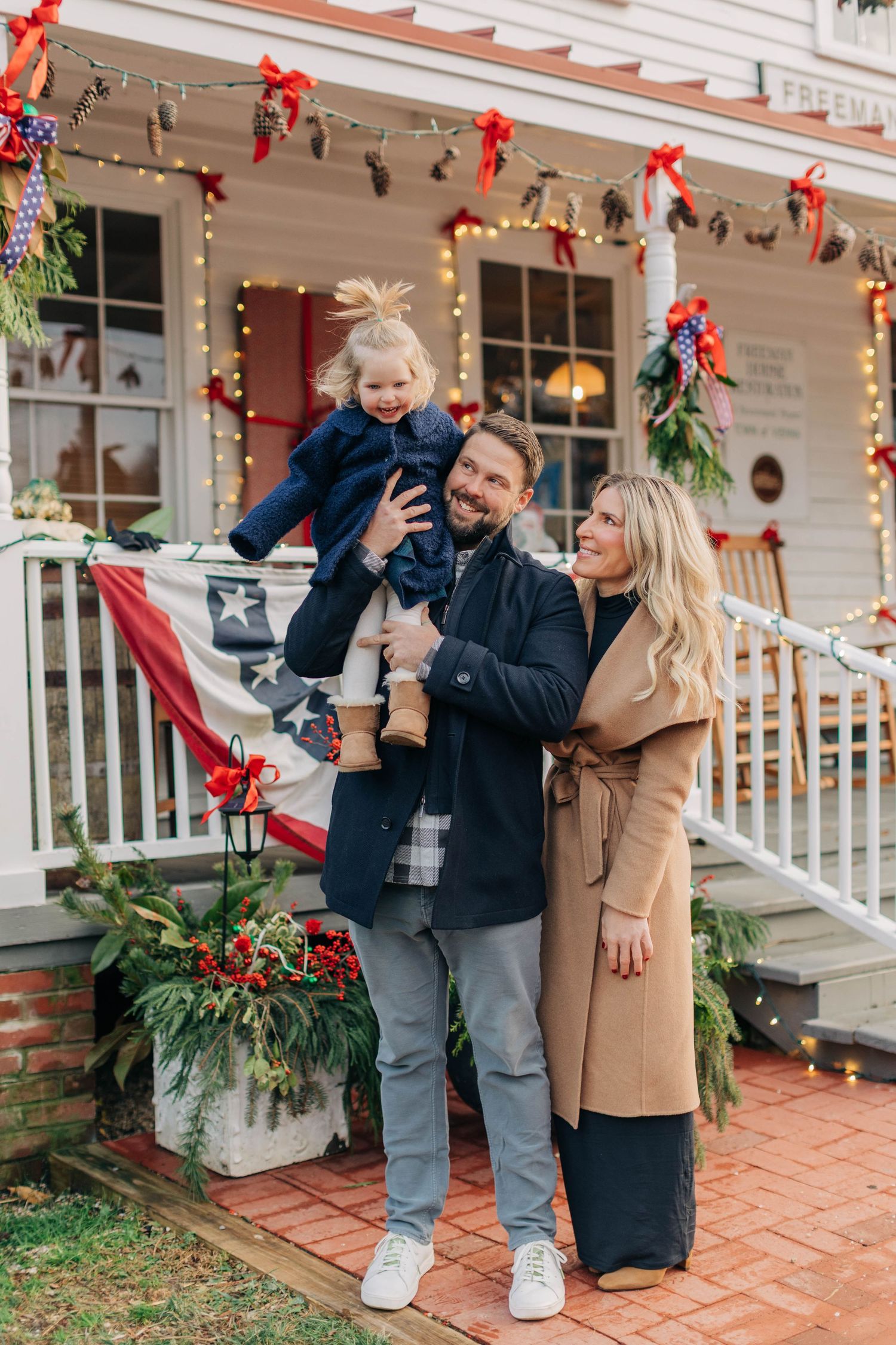 Happy family posing outdoors during Christmas, man holding toddler girl, woman in camel coat, festive decorated porch behind them.