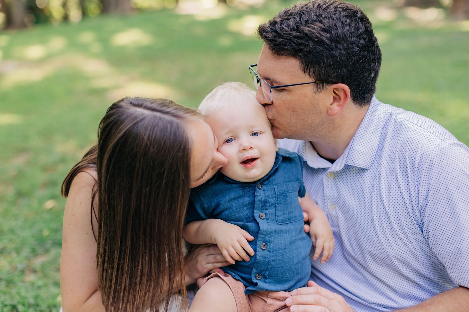 Parents kissing their smiling baby on the cheeks while sitting on green grass outdoors.